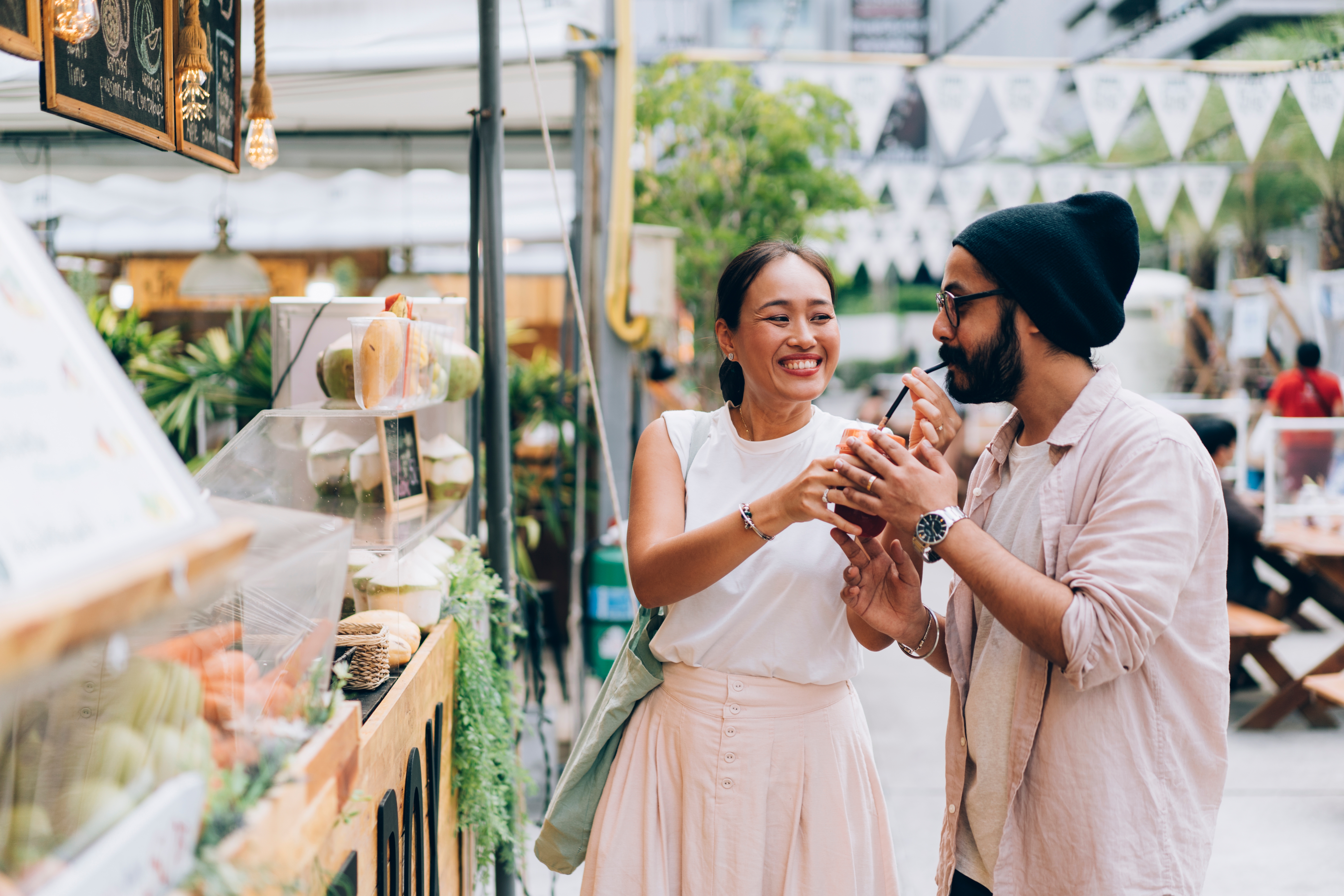 A smiling couple shares a playful moment in an outdoor market, with the woman playfully holding a snack. They seem happy and engaged with each other