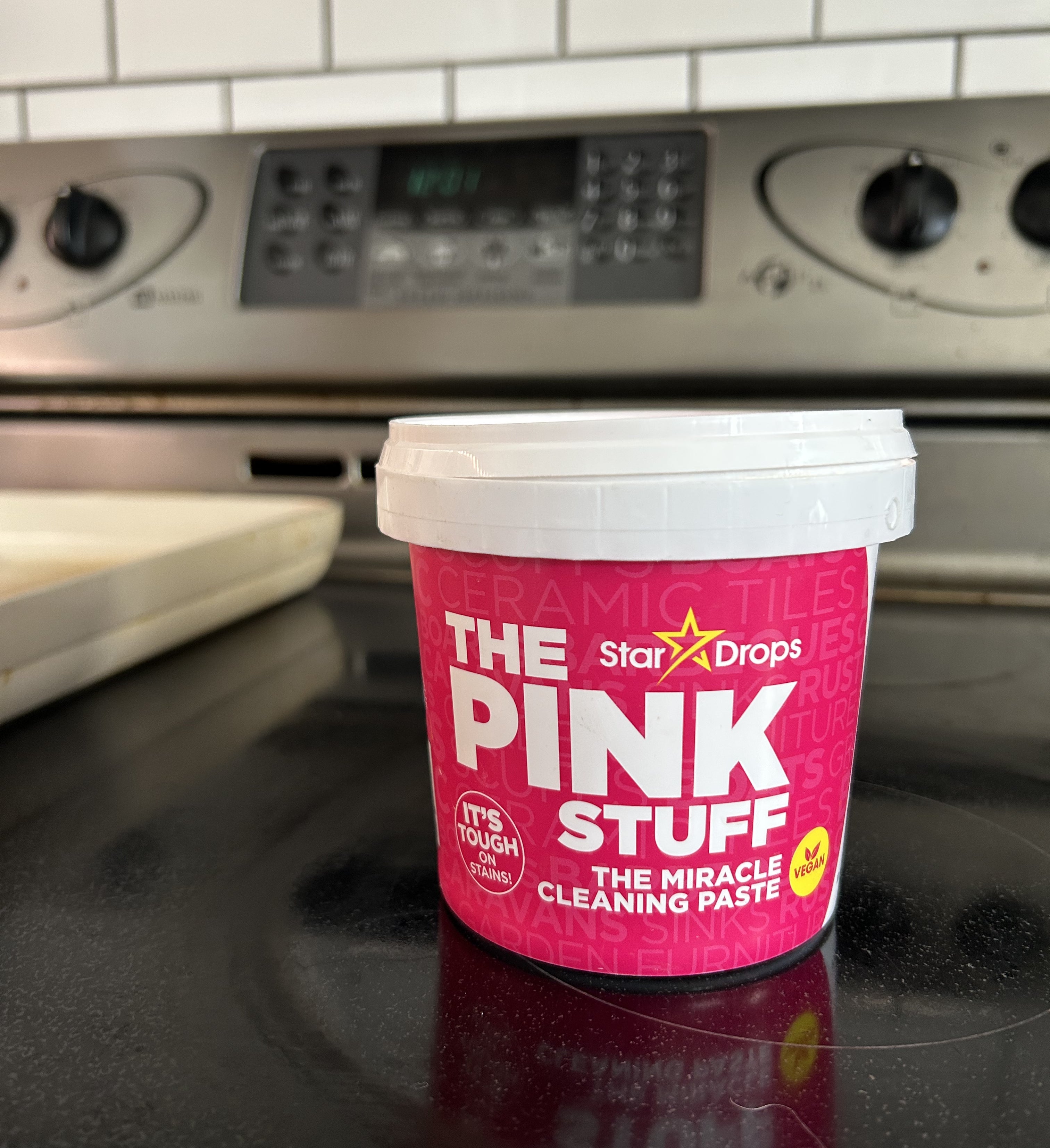 Container of The Pink Stuff cleaning paste on a stovetop, with kitchen tiles and an oven in the background