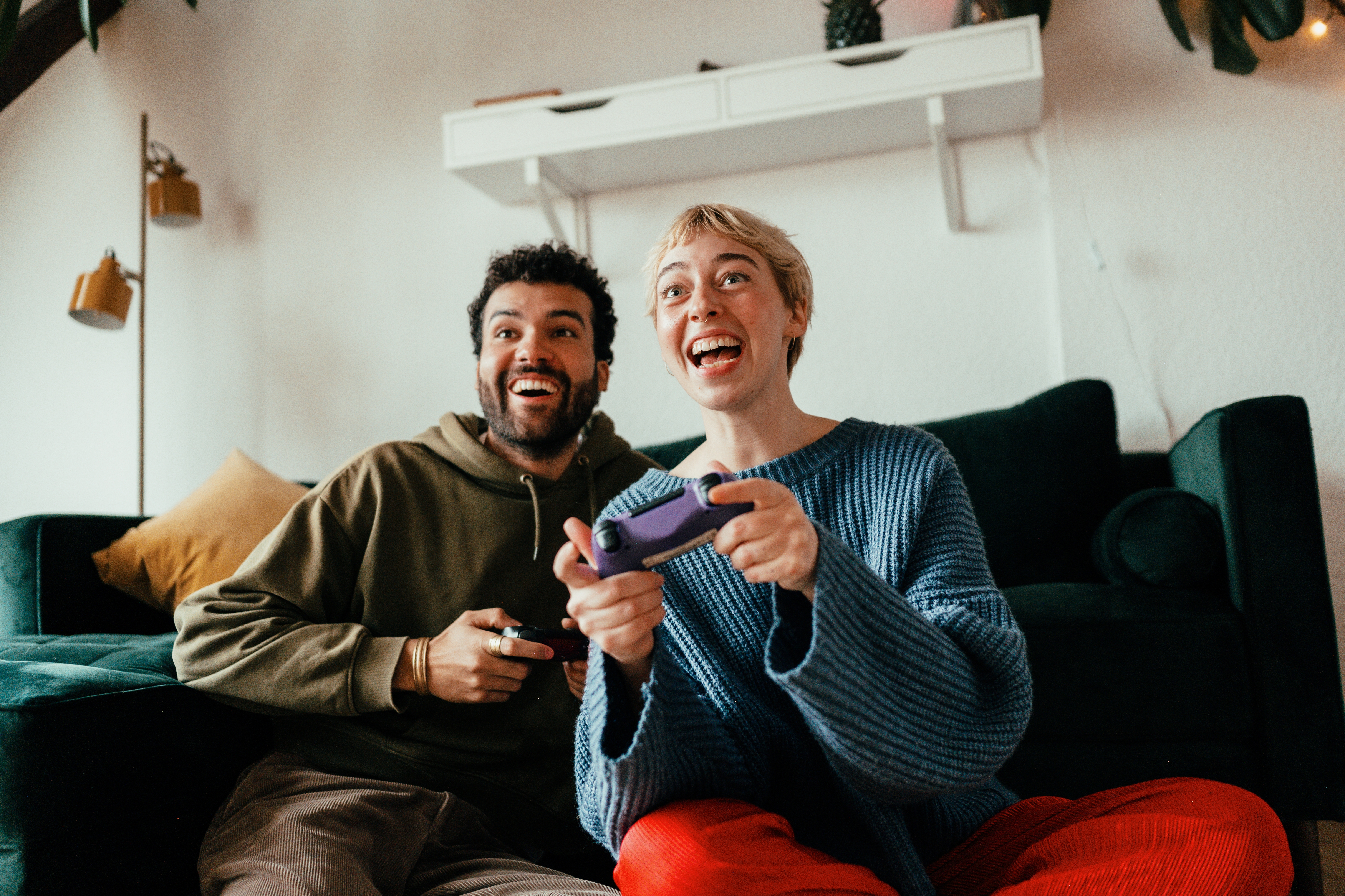 Two people sitting on a couch, joyfully playing video games with controllers, one wearing a cozy sweater and the other in a casual hoodie
