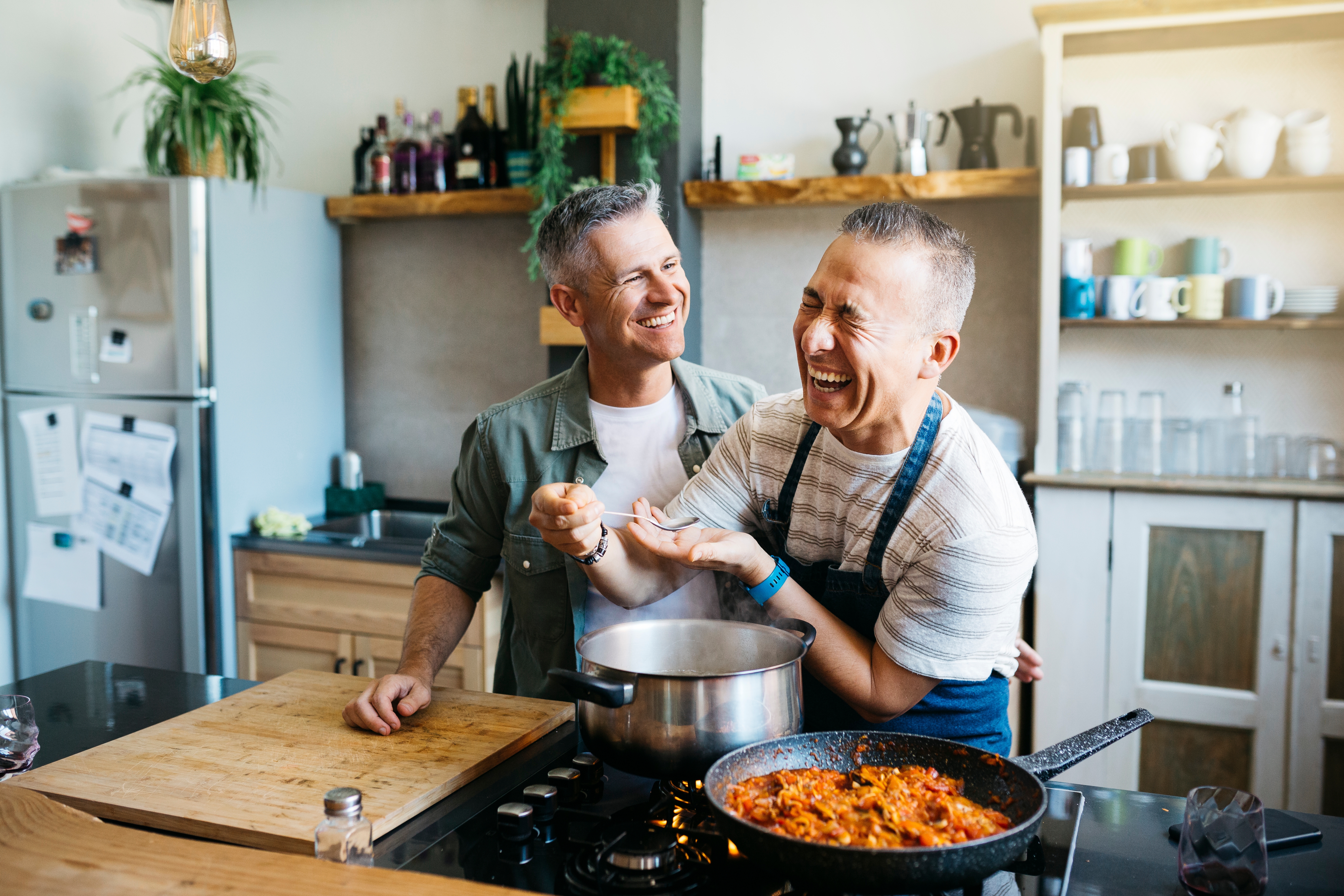 Two people in a kitchen, smiling and cooking together, enjoying a joyful moment while preparing a meal on the stove
