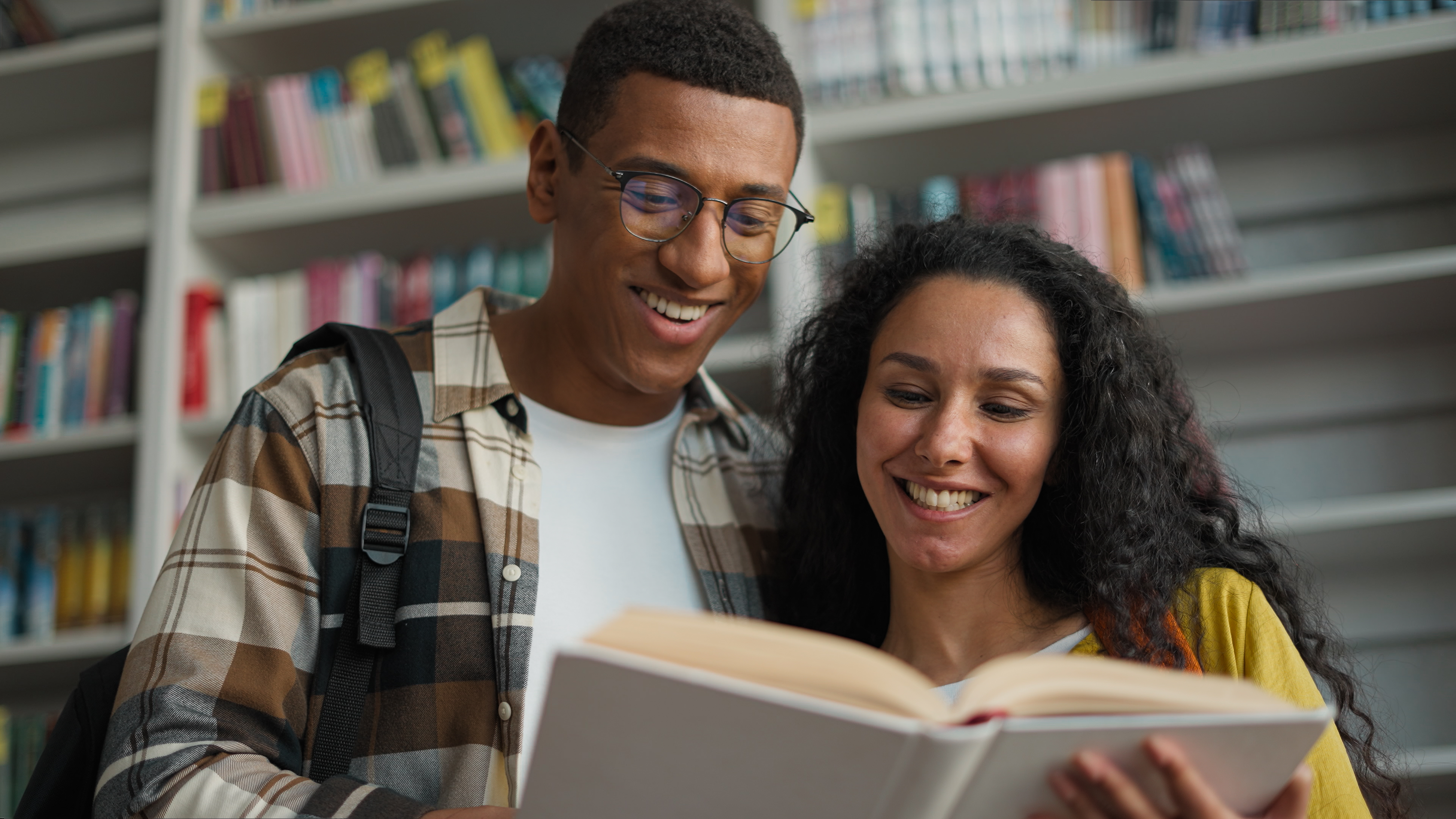 Two people smiling and reading a book together in a library, conveying a sense of shared interest and intimacy