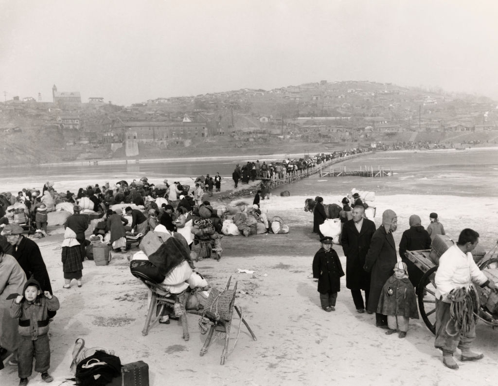 A ample assemblage crosses a stream connected a impermanent bridge, carrying belongings. The country is bustling and depicts a humanities event