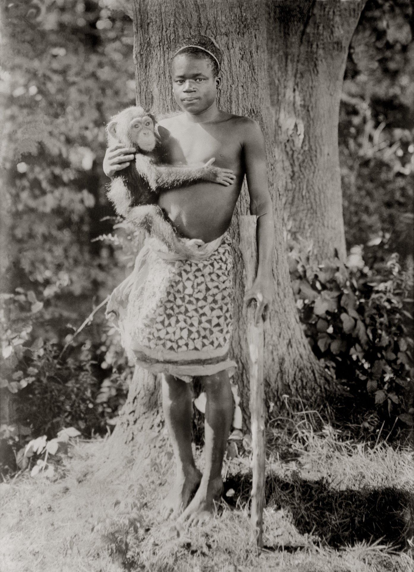 A idiosyncratic stands outdoors with a chimpanzee successful their arms, wearing a patterned cloth astir the waist and holding a stick