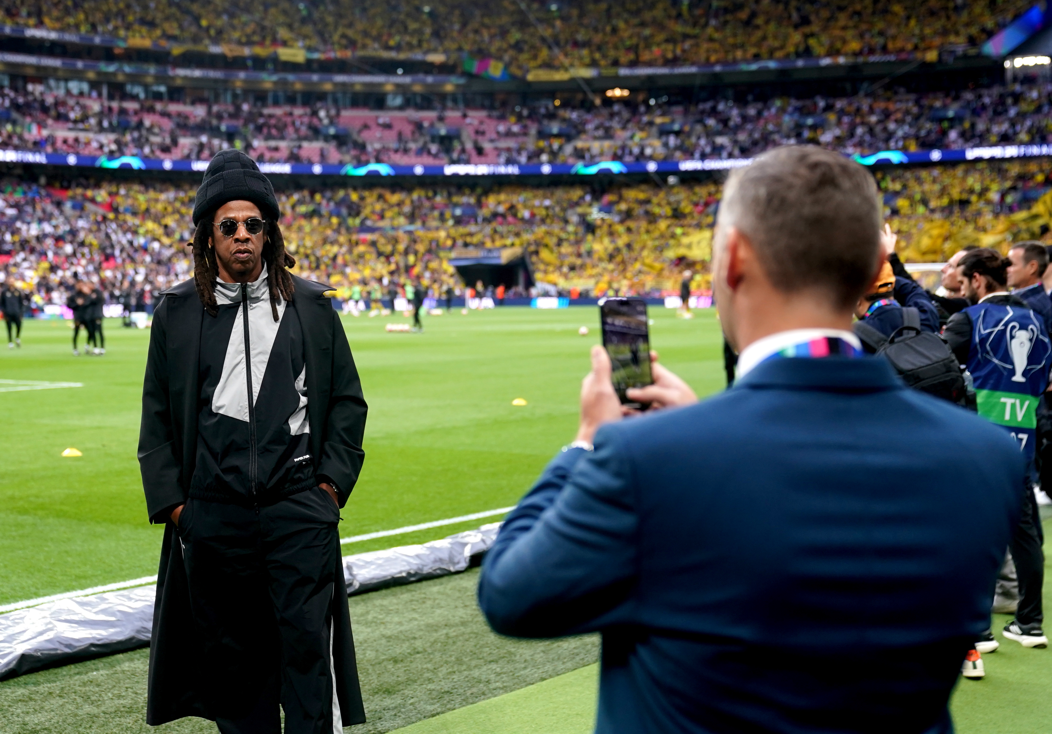 Celebrity wearing agelong  overgarment  and chapeau  astatine  sports event, being photographed by different  idiosyncratic   connected  the field. Stadium assemblage  disposable   successful  the background