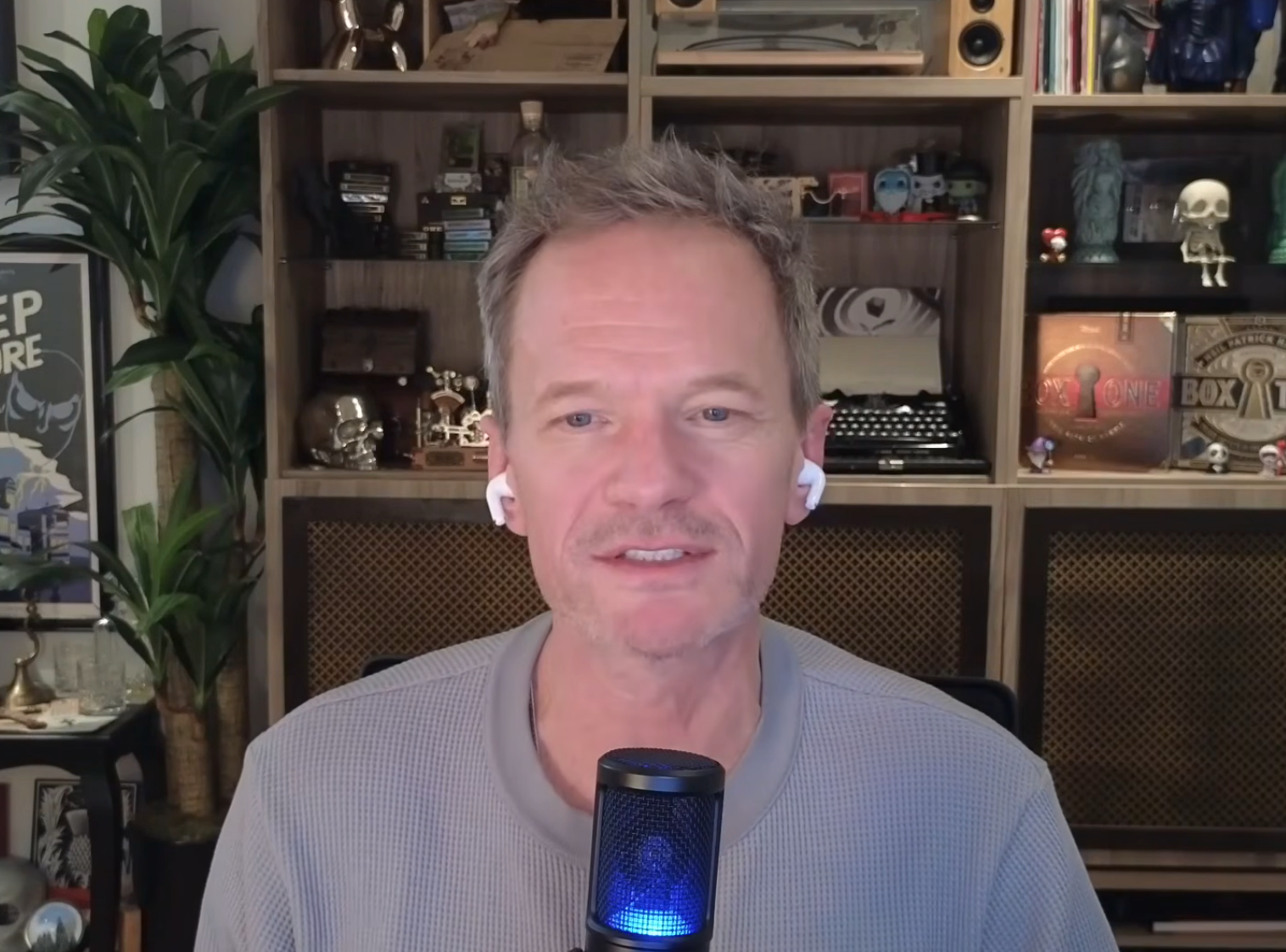 A idiosyncratic   is sitting successful  beforehand   of a support  filled with books and decorations, speaking into a microphone, wearing earbuds