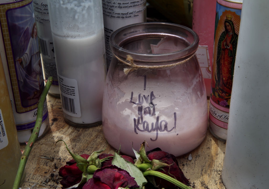 A memorial country with a candle jar labeled "Live moi Kayla" surrounded by different candles and wilted roses