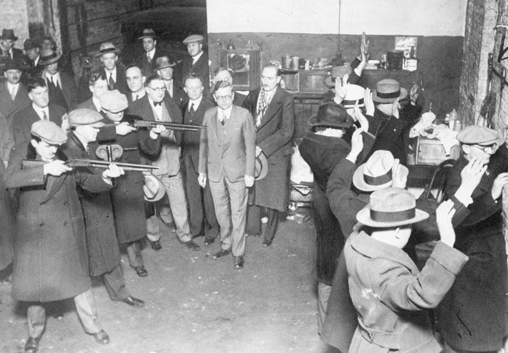 Historical reenactment of a prohibition-era scene, showing men successful suits and fedoras with hands raised, surrounded by men with guns