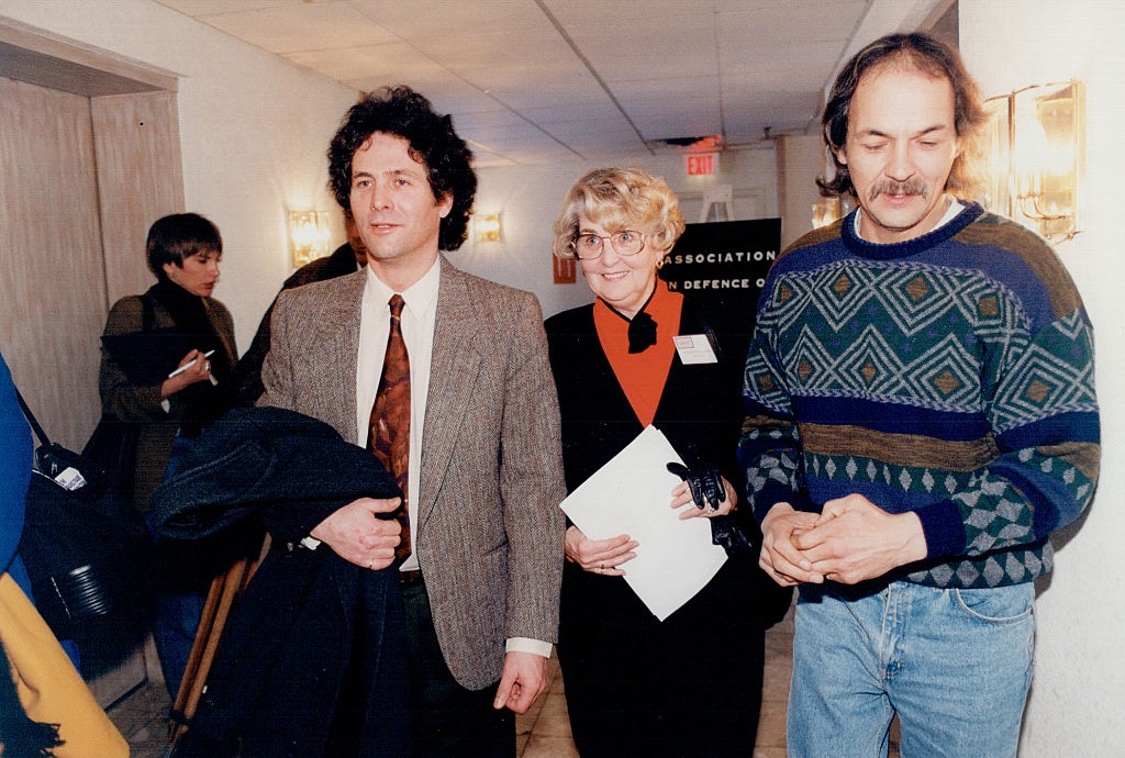 Three radical walking indoors; 2 men, 1 with a suit and different with a patterned sweater, and a pistillate successful glasses holding papers