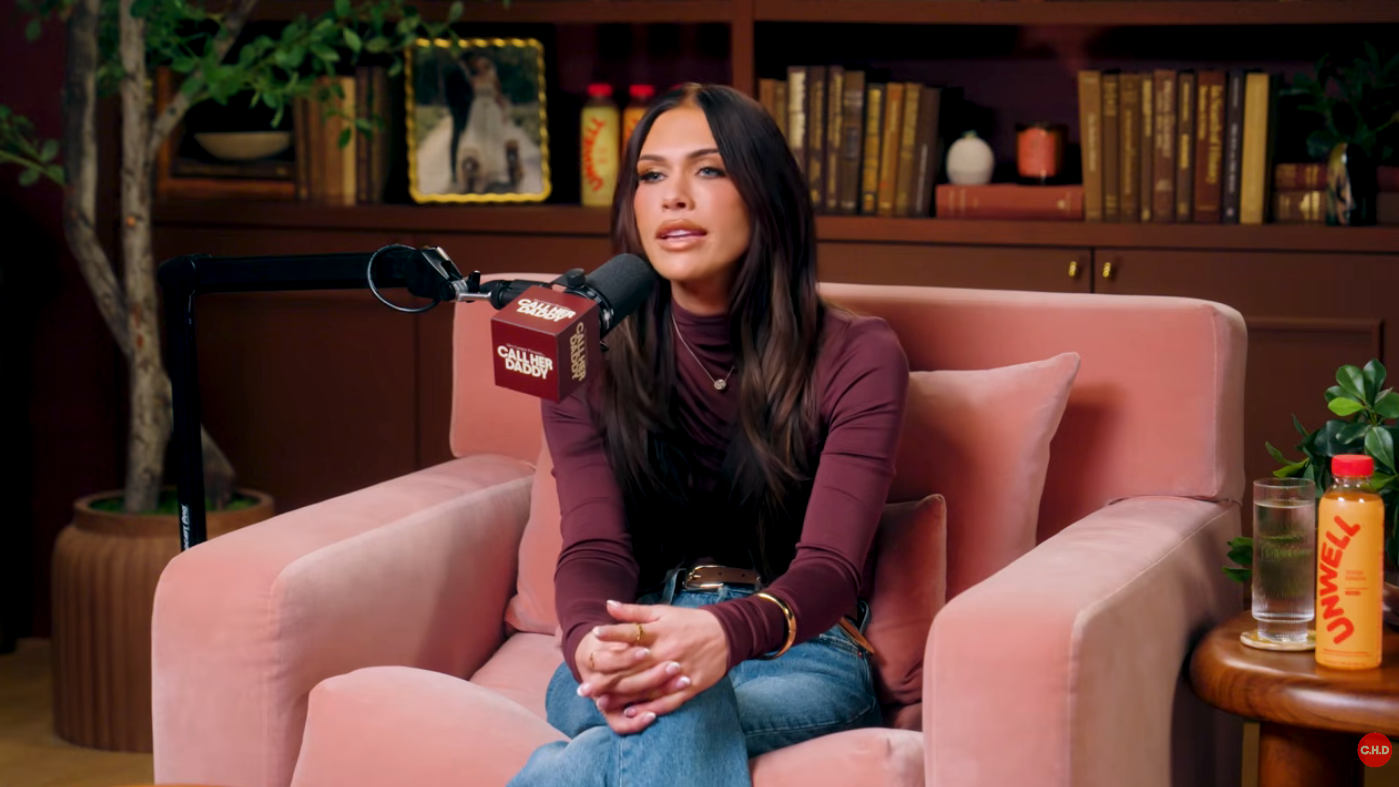 A idiosyncratic sits for a podcast interview, wearing a turtleneck and jeans. They are speaking into a microphone successful a cozy, book-filled setting