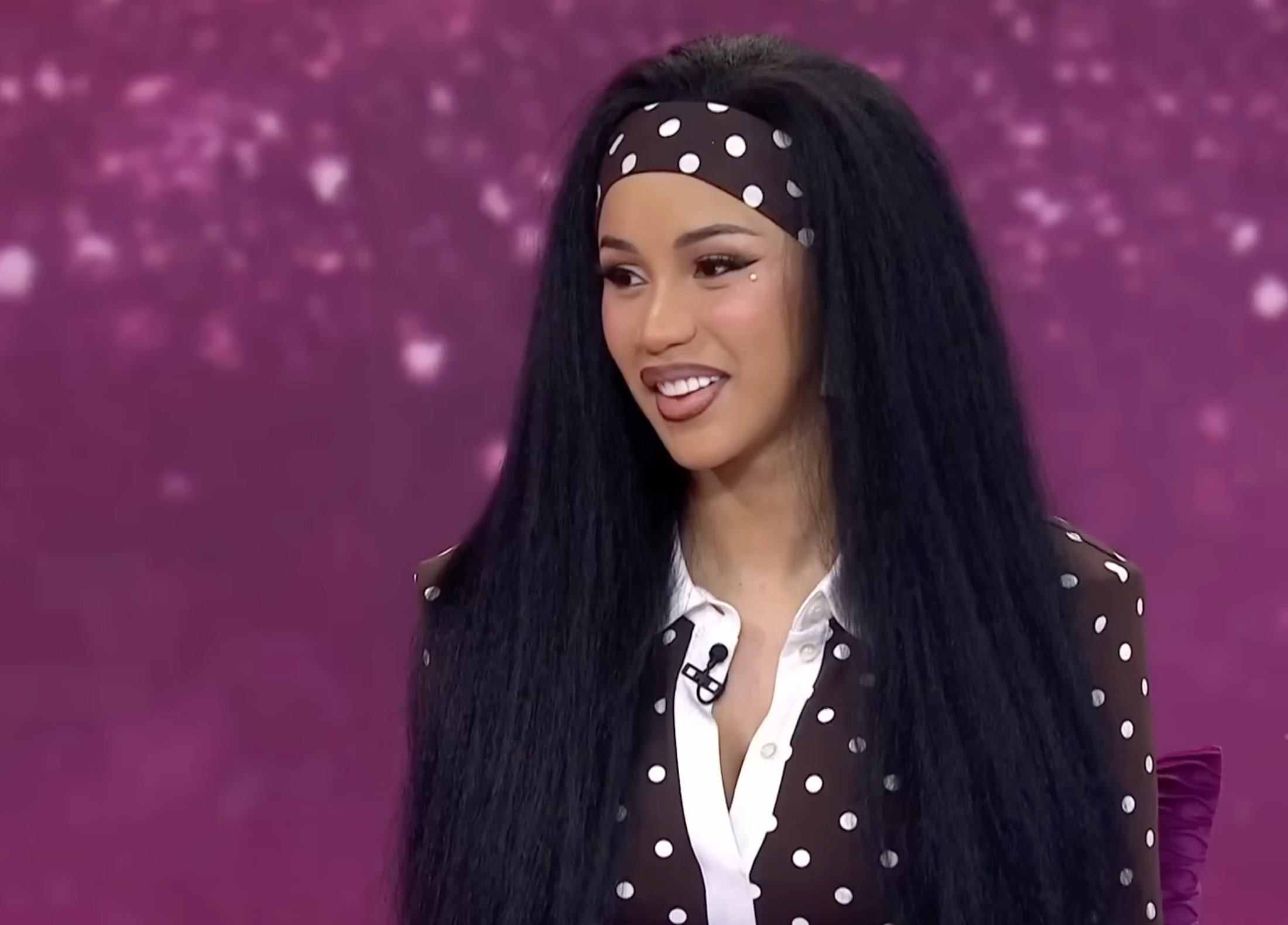 Person with long hair and polka dot headband, wearing a matching polka dot blouse, seated on a talk show set with a purple background