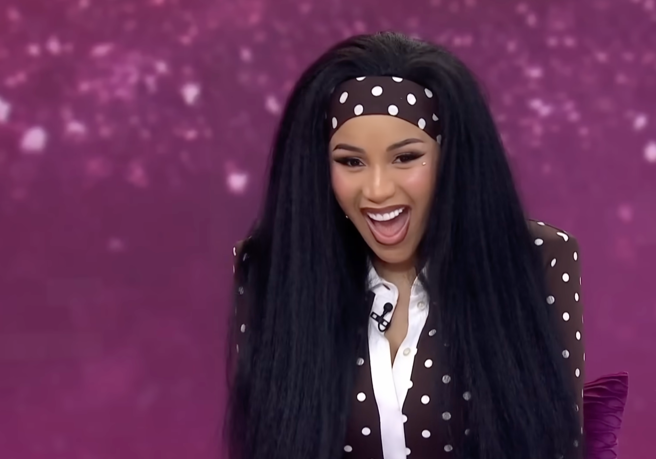 Person with long hair, wearing a polka dot outfit and matching headband, smiling joyfully during an on-stage appearance