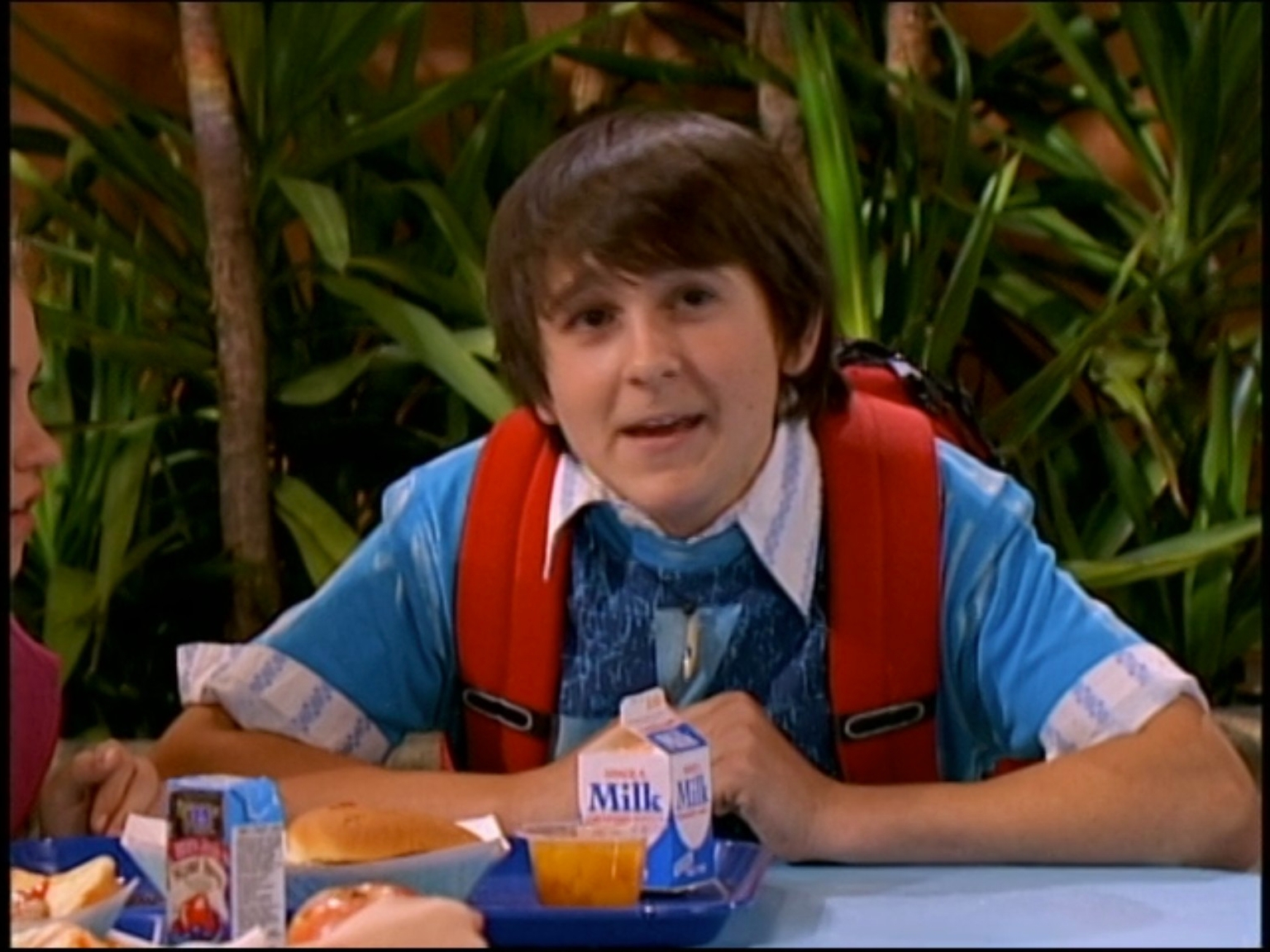 A young idiosyncratic with a shaggy hairstyle sits astatine a cafeteria array with a nutrient tray, wearing a backpack implicit a casual shirt