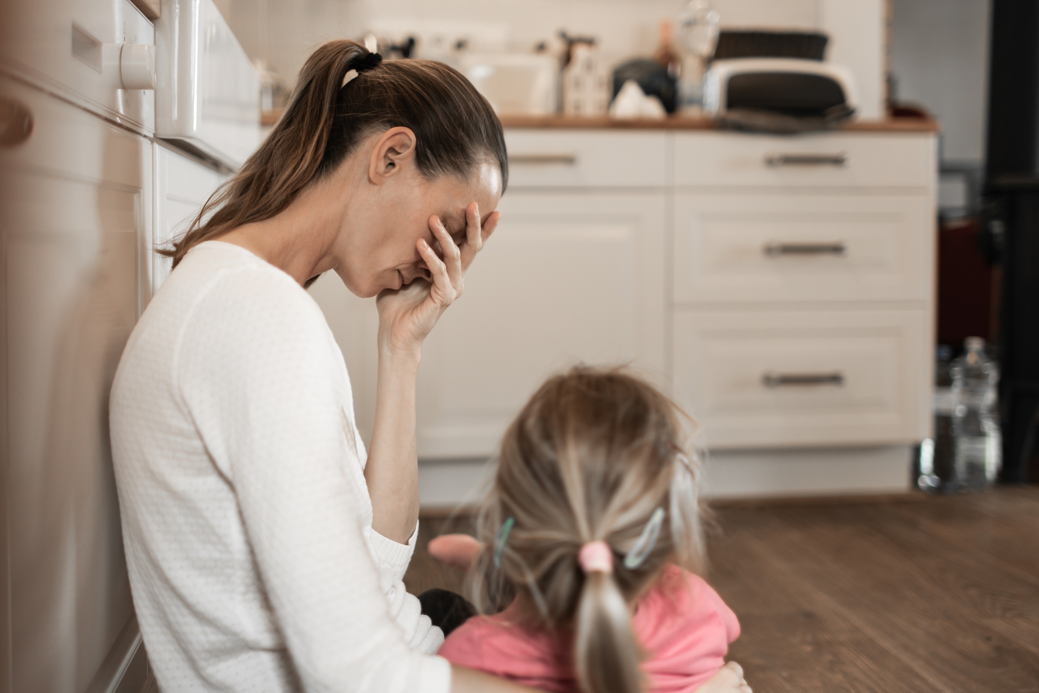 A woman sits on a kitchen floor, covering her face with her hand, as a young child sits nearby facing her