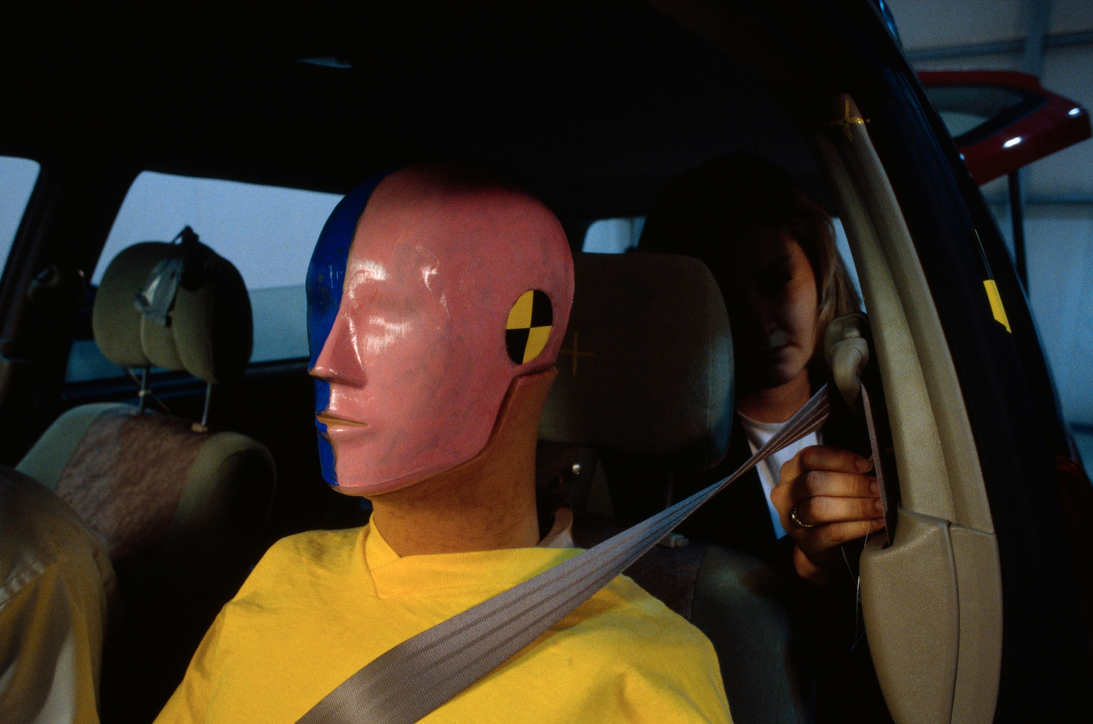 A crash test dummy is seated in a car while a person buckles the seatbelt, demonstrating vehicle safety measures