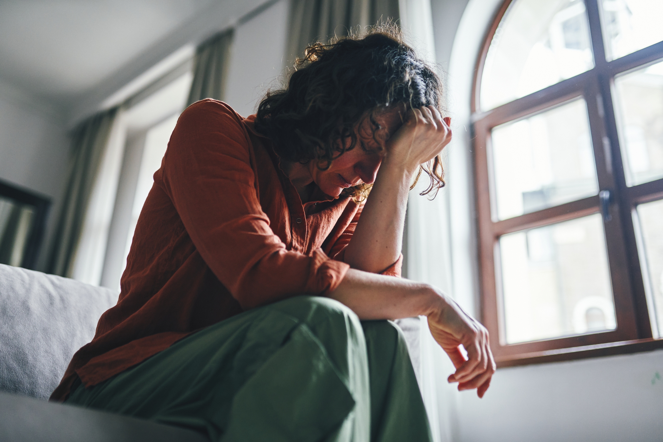 Person sitting on a couch, looking distressed with head in hand, near a window