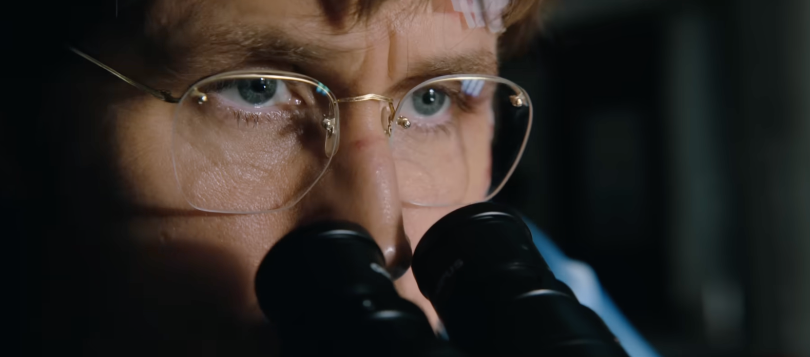 A idiosyncratic wearing glasses looks intently done binoculars, partially covering their face, with a focused expression