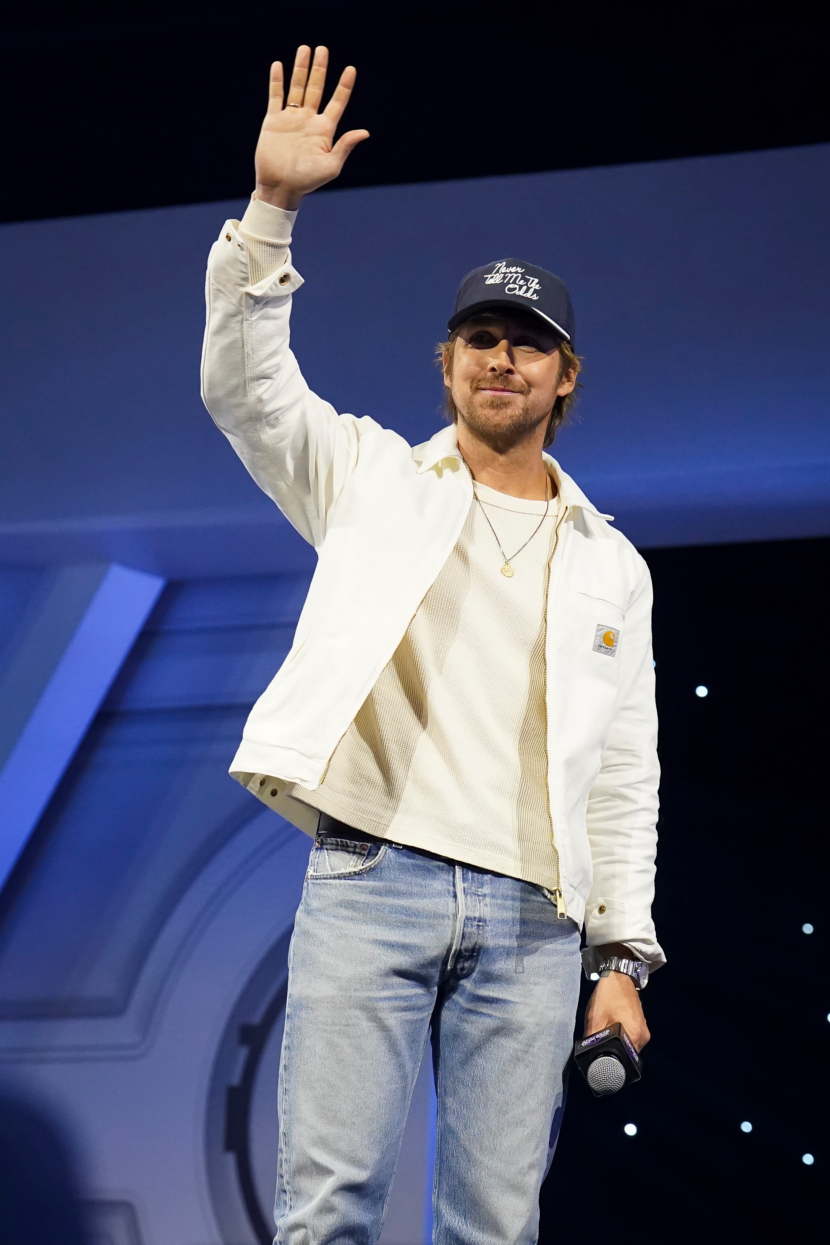Person connected signifier wearing casual jeans, a achromatic jacket, and a cap, holding a microphone, waves to an audience