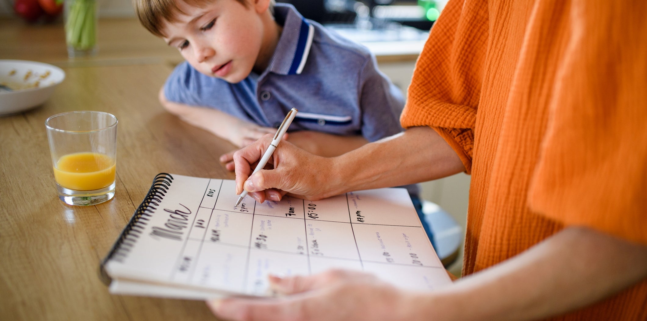 A person fills out a March calendar on a kitchen counter with a curious child looking on, next to a glass of orange juice