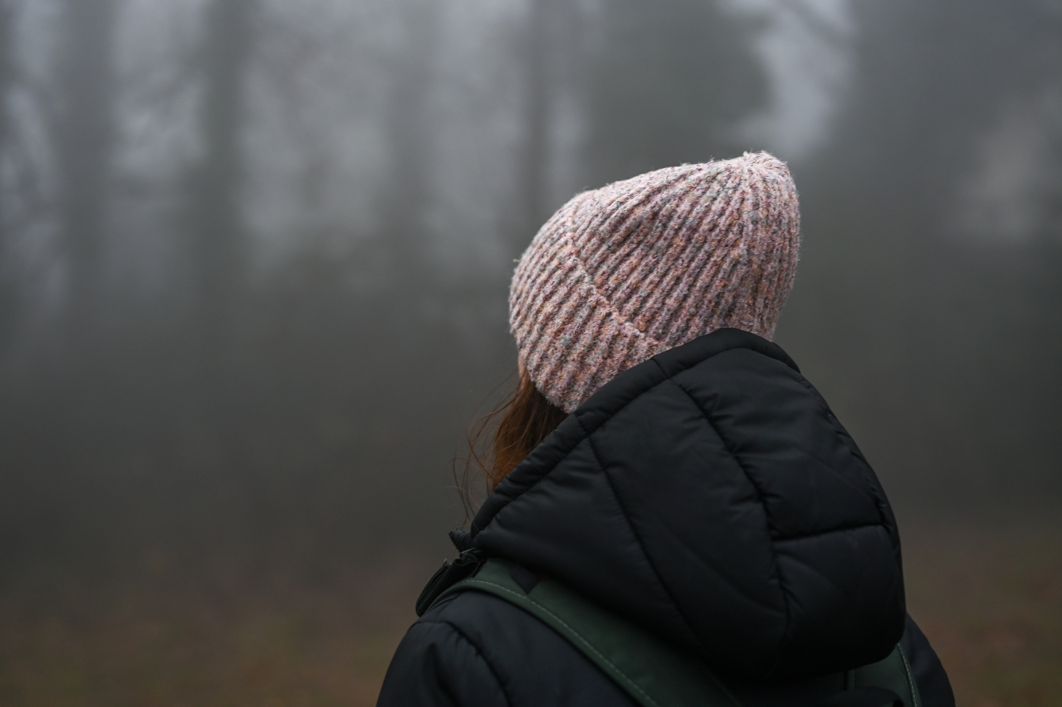 Person in a cozy knit hat and jacket stands alone, gazing into a misty, wooded landscape, evoking a sense of solitude and introspection