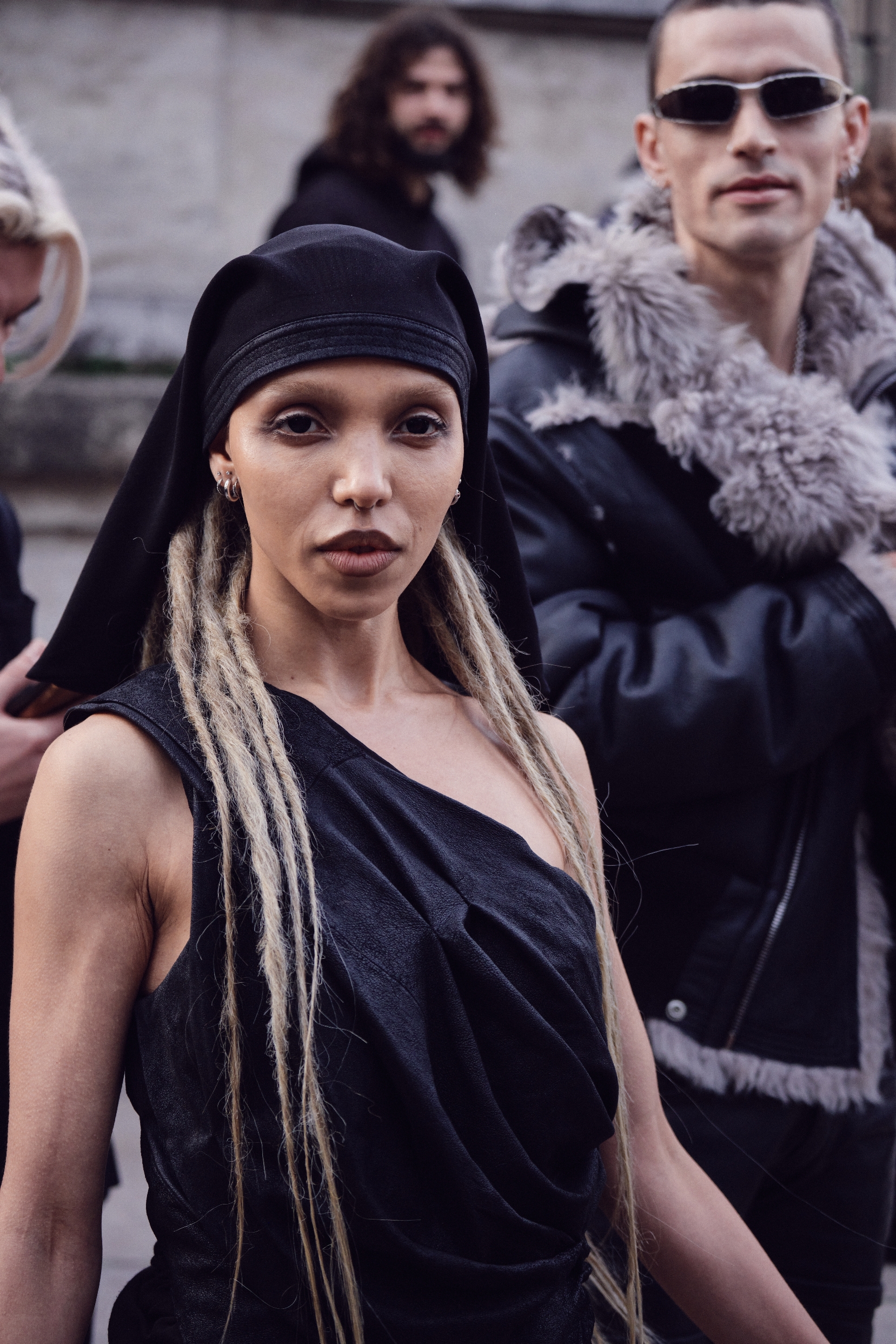 A idiosyncratic   successful  a draped, one-shoulder outfit with agelong  braids and a headscarf poses confidently. Another idiosyncratic   successful  sunglasses and a fur-trimmed overgarment   stands nearby