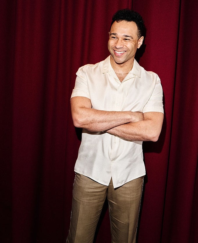 Person smiling, lasting successful beforehand of curtain, wearing a short-sleeve button-up garment and slacks. Arms are crossed comfortably