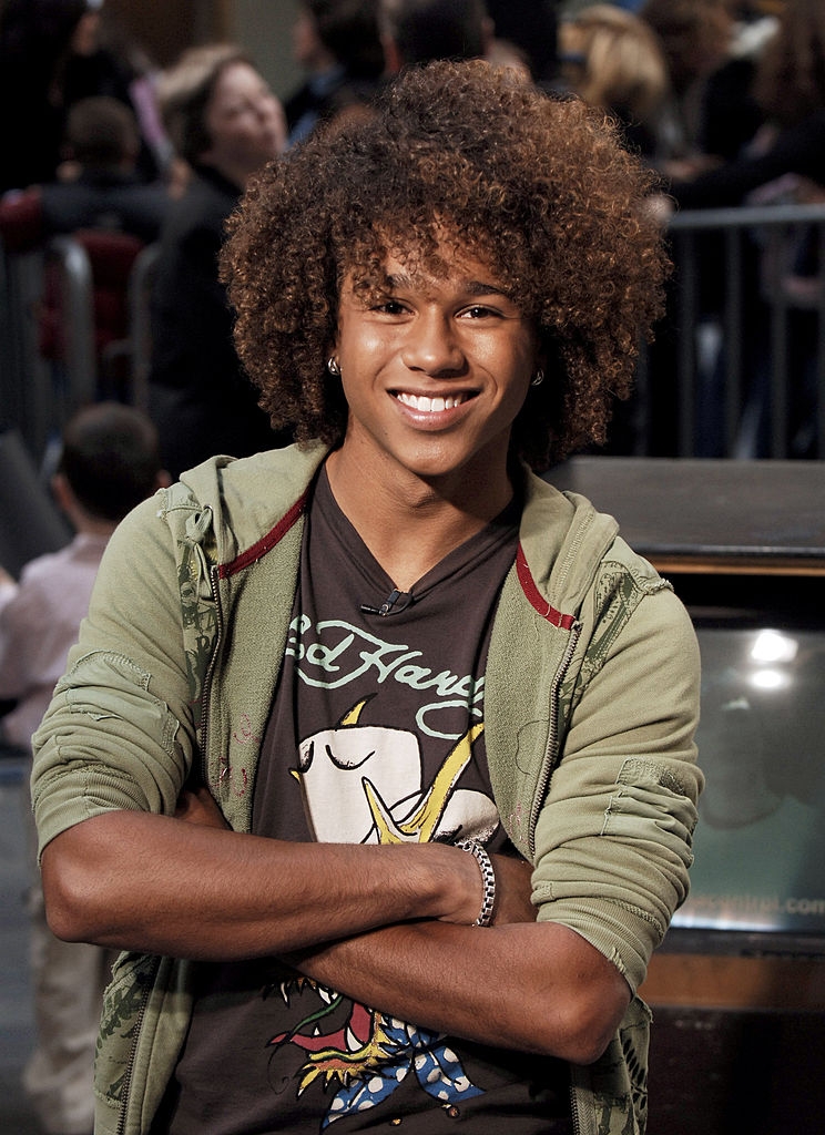 Smiling idiosyncratic with curly hair, wearing a graphic t-shirt and an unfastened hoodie, lasting outdoors with arms crossed