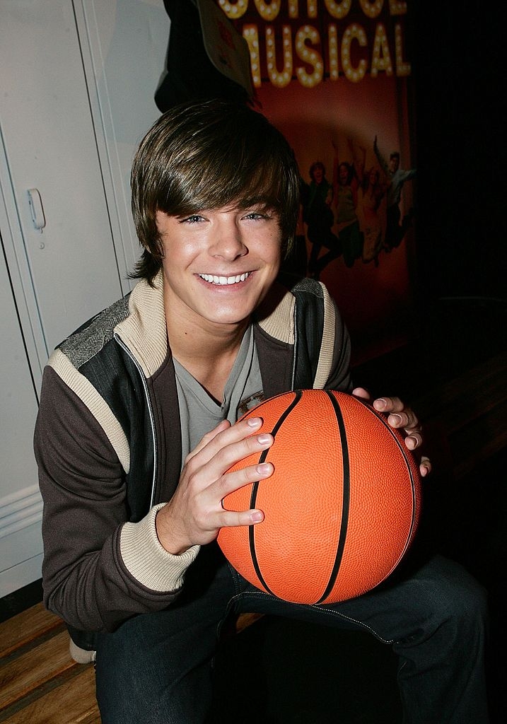 Person smiling, holding basketball, successful beforehand of "School Musical" sign. Wearing casual layered clothing