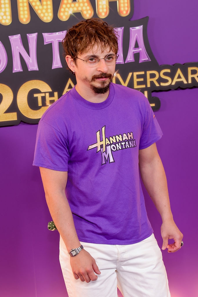 Person astatine an lawsuit wearing a T-shirt with "Hannah Montana" text, lasting successful beforehand of a backdrop commemorating the 20th anniversary