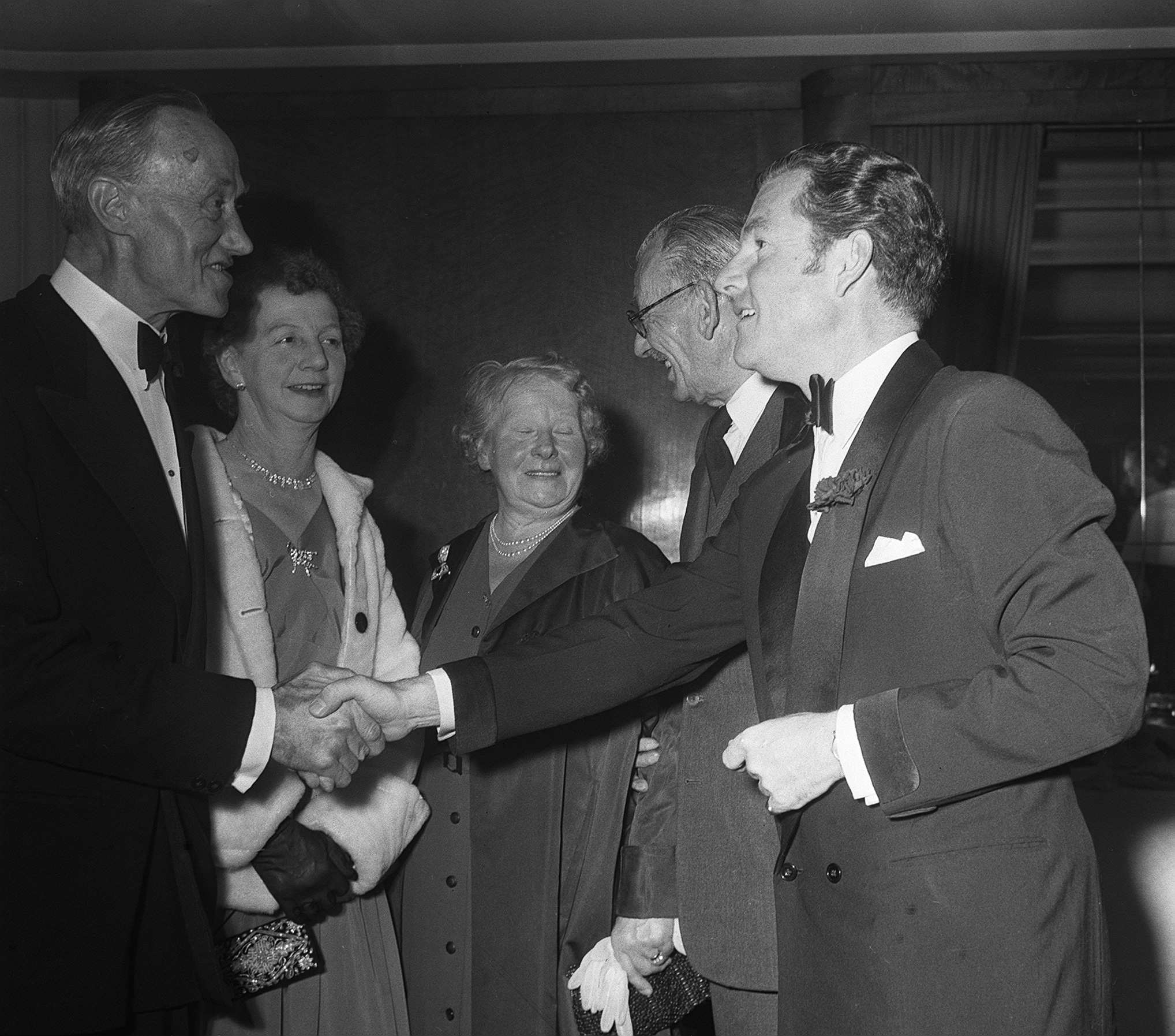 A radical of elegantly dressed radical astatine a ceremonial event, 2 men successful tuxedos shingle hands warmly portion others look on, smiling