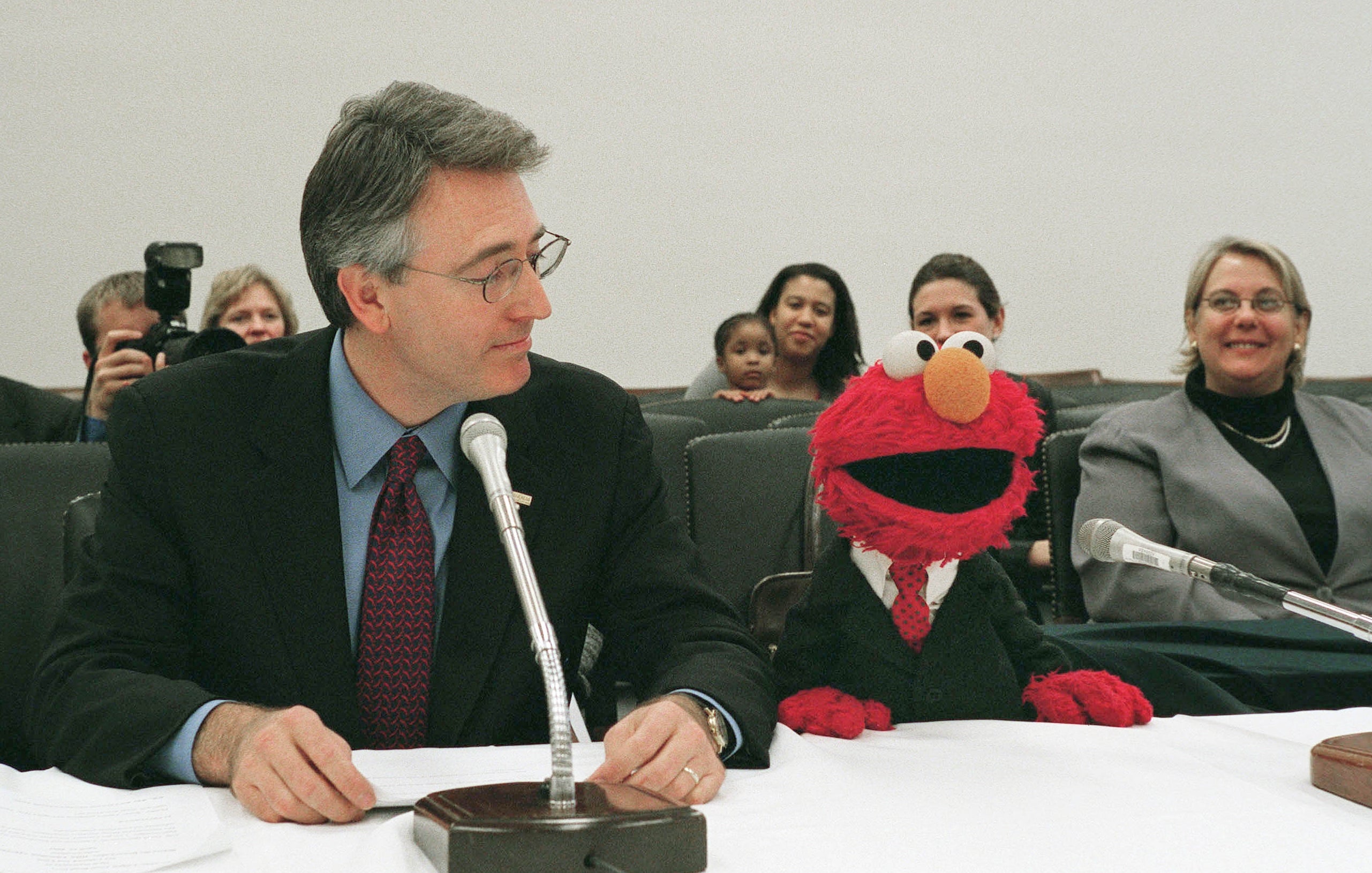 Elmo, dressed successful a suit, sits astatine a array with a antheral successful a suit, speaking into microphones astatine a nationalist event