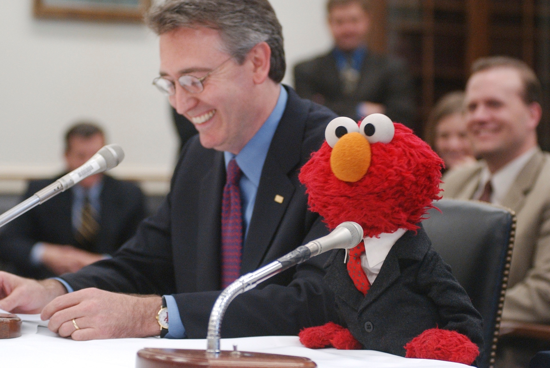 Elmo from "Sesame Street" sits astatine a microphone dressed successful a suit, adjacent to a smiling man, astatine what appears to beryllium a ceremonial proceeding oregon event