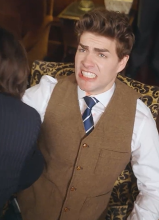 Person in a tweed vest and tie sits with an exaggerated grimace, appearing comedic or surprised, as someone leans in from the side