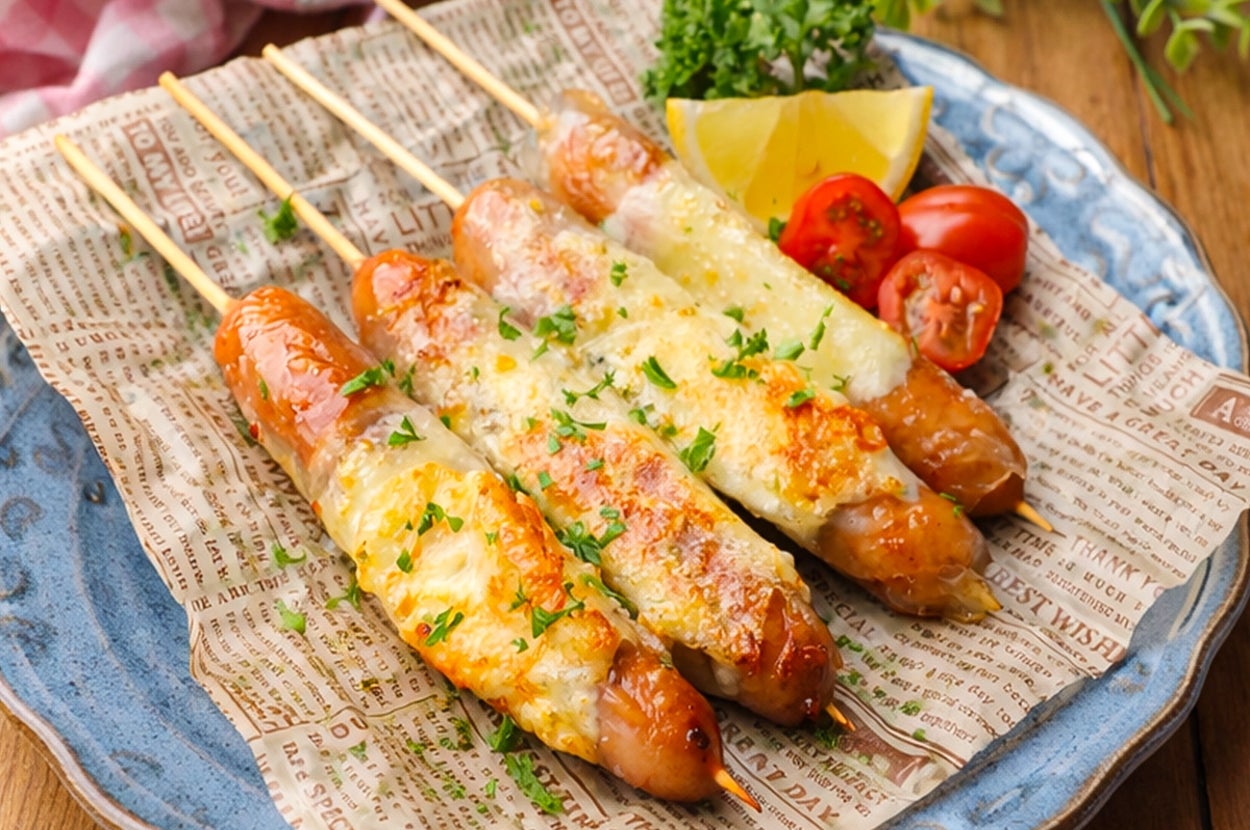 お皿に並んだチーズがけソーセージの串焼き。トマト、レモン、ハーブが添えられている。