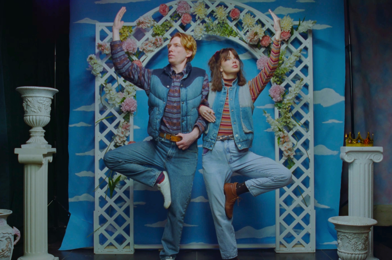 Taylor Swift and Domhall Gleeson pose playfully in denim outfits, in front of a sky-themed backdrop with a floral arch in "Opalite" music video
