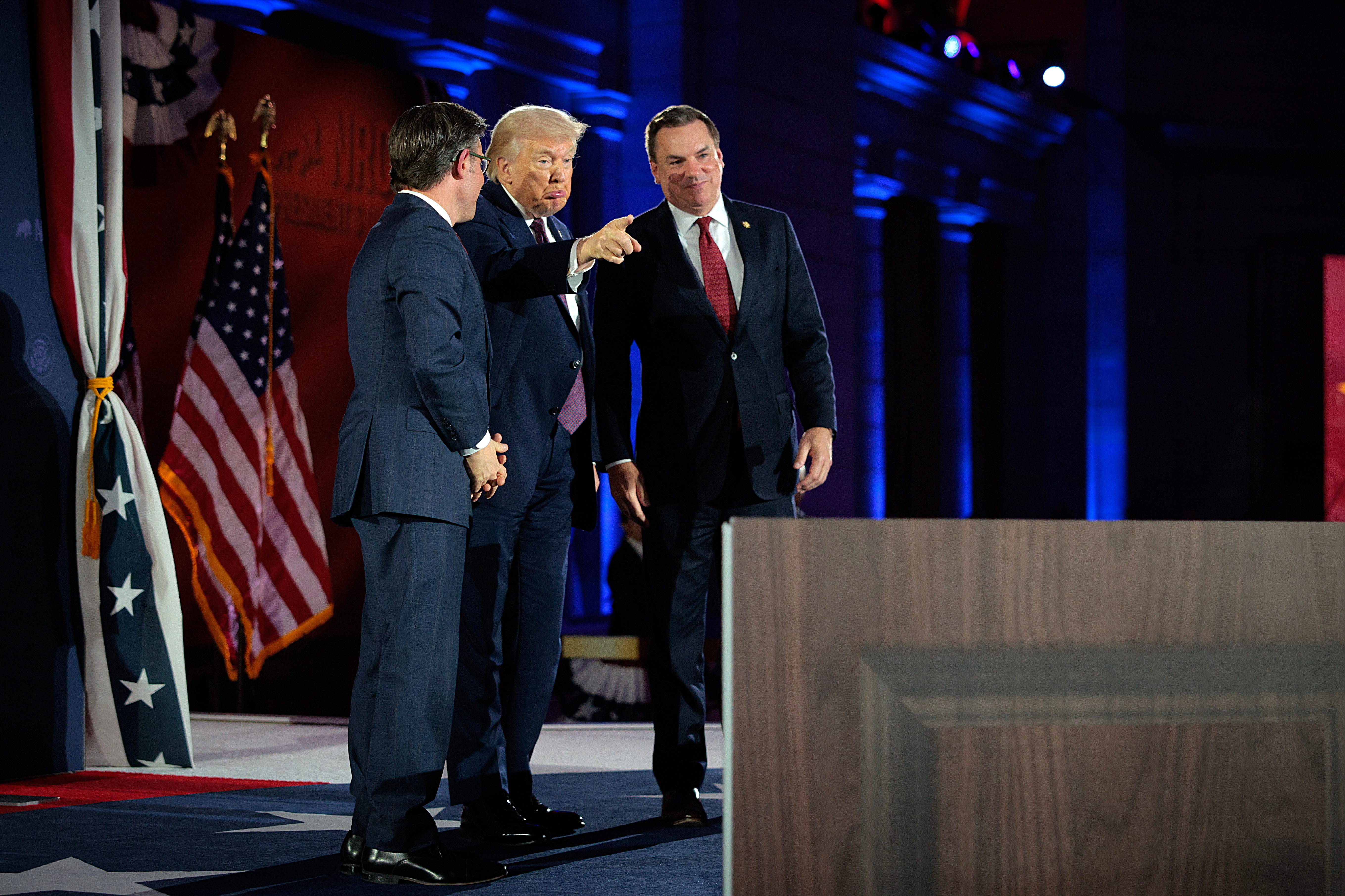 Three men successful suits prosecute successful speech connected a signifier with flags successful the background. One points forward, indicating thing off-frame