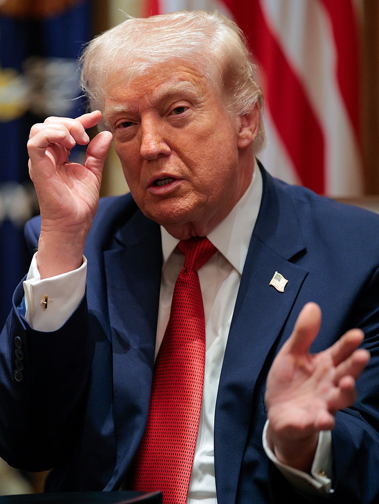 Person successful a suit gestures portion speaking, seated indoors with a emblem successful the background