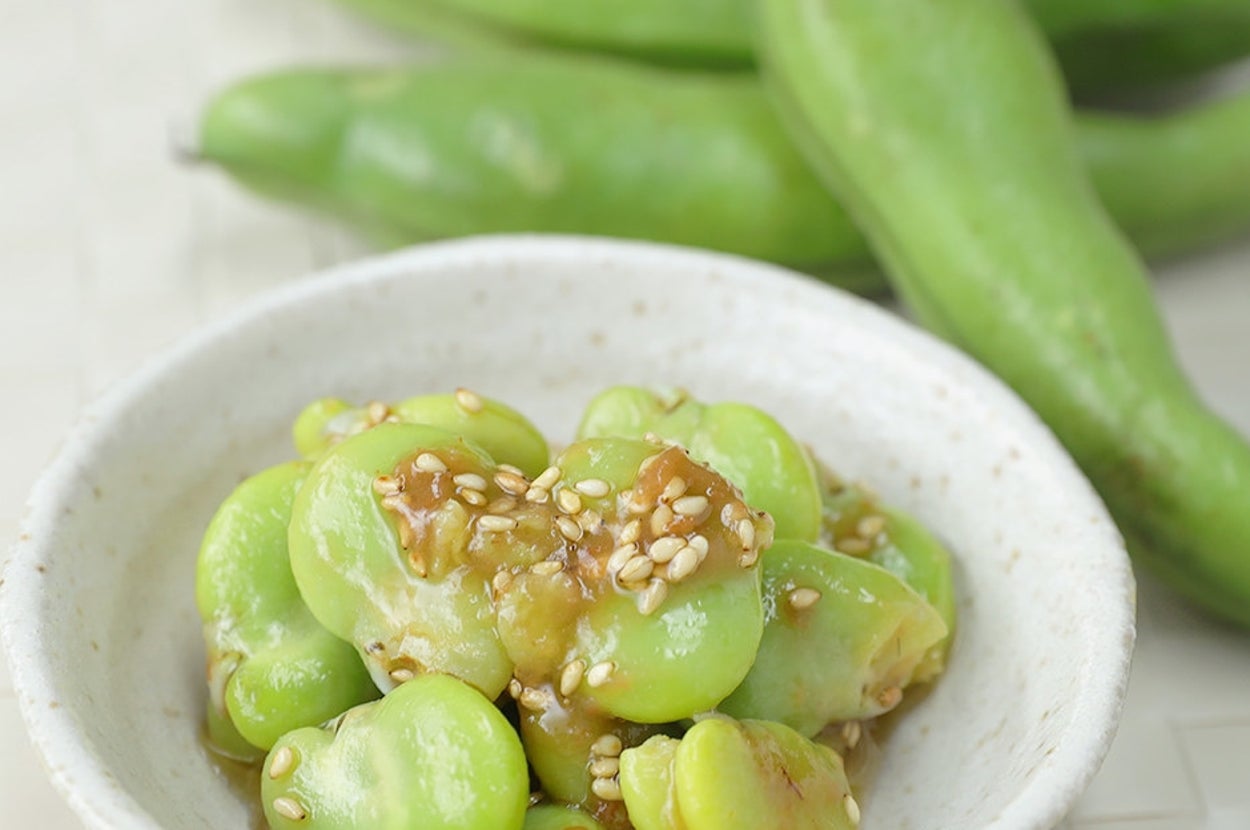 白い器に盛られたそら豆の和え物。器の後ろには、生のそら豆のさやが置かれている。