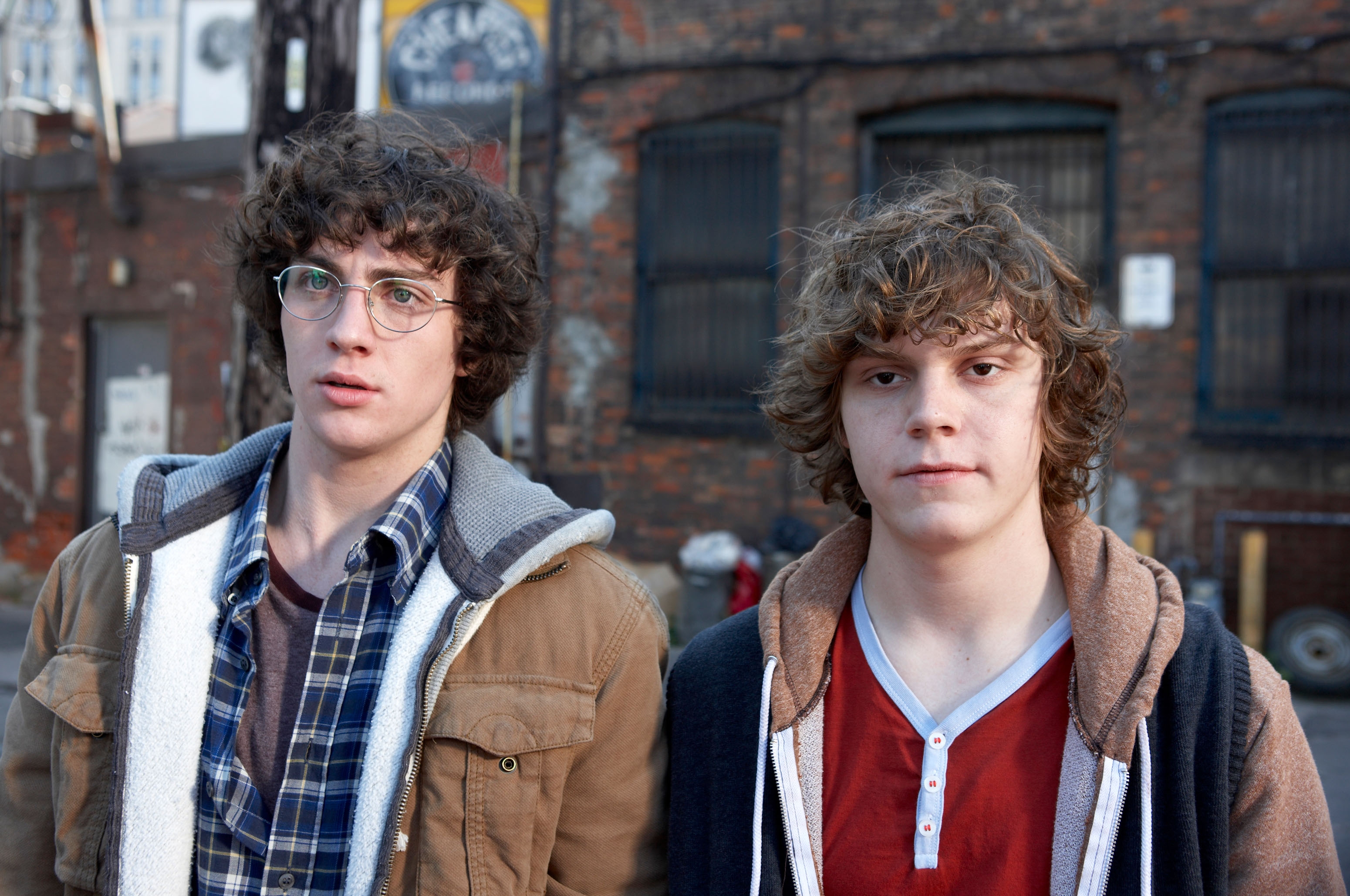 1  wears a overgarment   and glasses, the different   a hoodie with a reddish  garment  underneath. Urban background