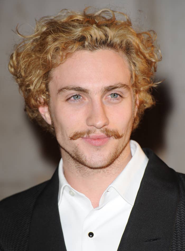 Person with curly hairsbreadth  and mustache, wearing a suit   with a achromatic  shirt, smiles slightly, looking distant  from the camera