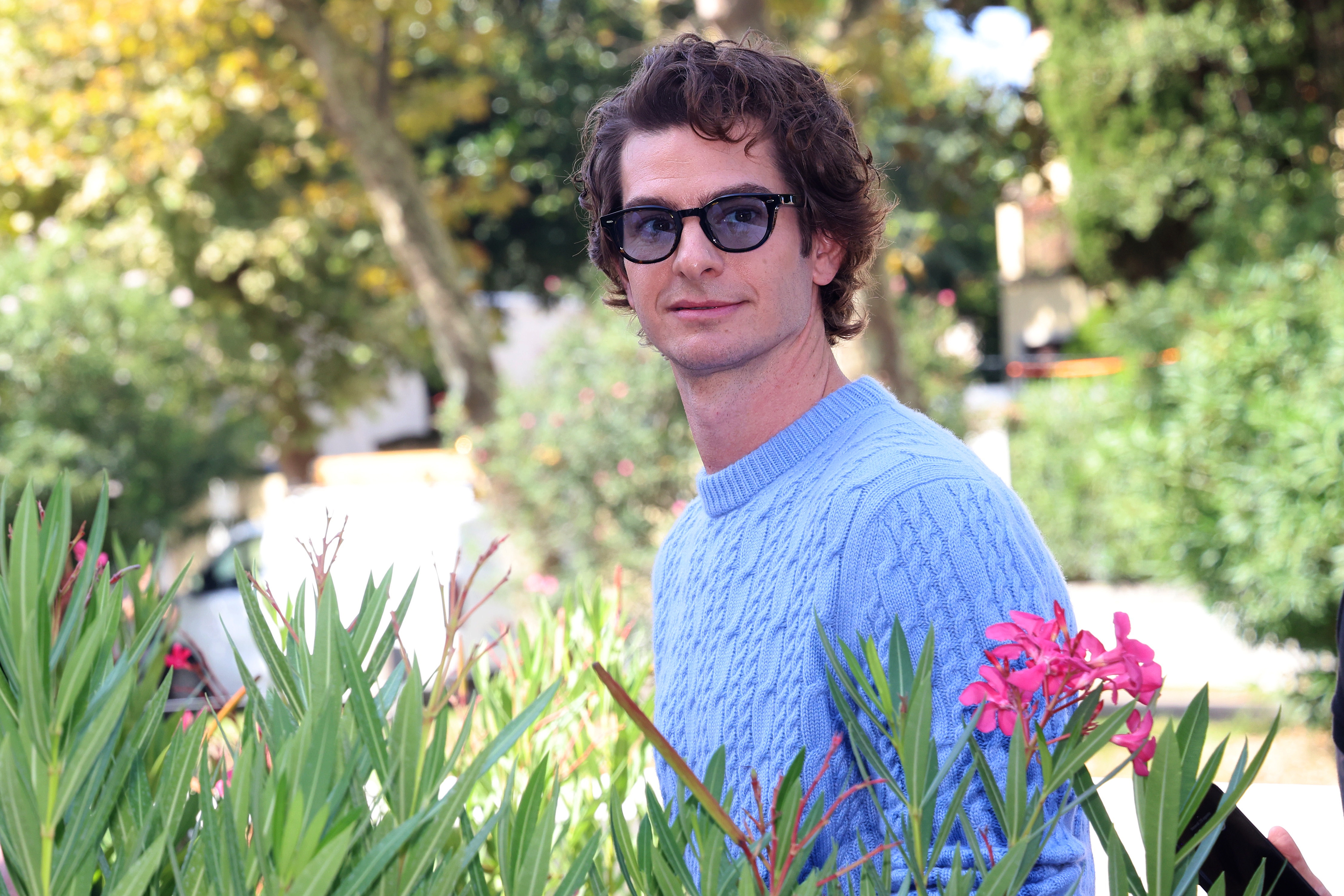 Person in a textured sweater and glasses standing outdoors among greenery and flowers