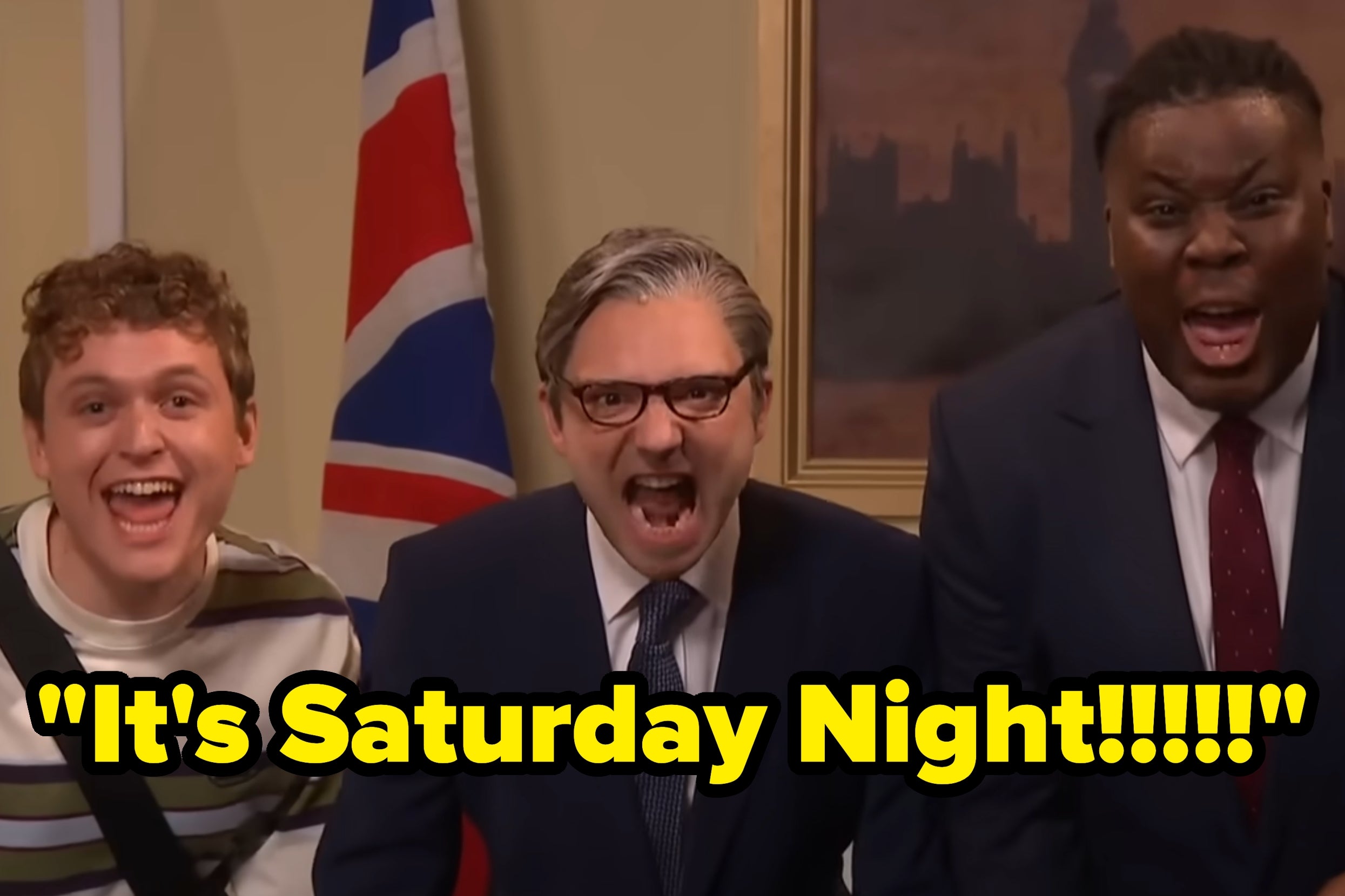 Three excited men in suits and casual wear shout "It's Saturday Night!!!!!!" near a British flag