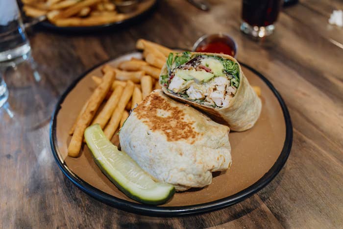 A plate with a halved wrap filled with chicken and greens, served with fries and a pickle spear