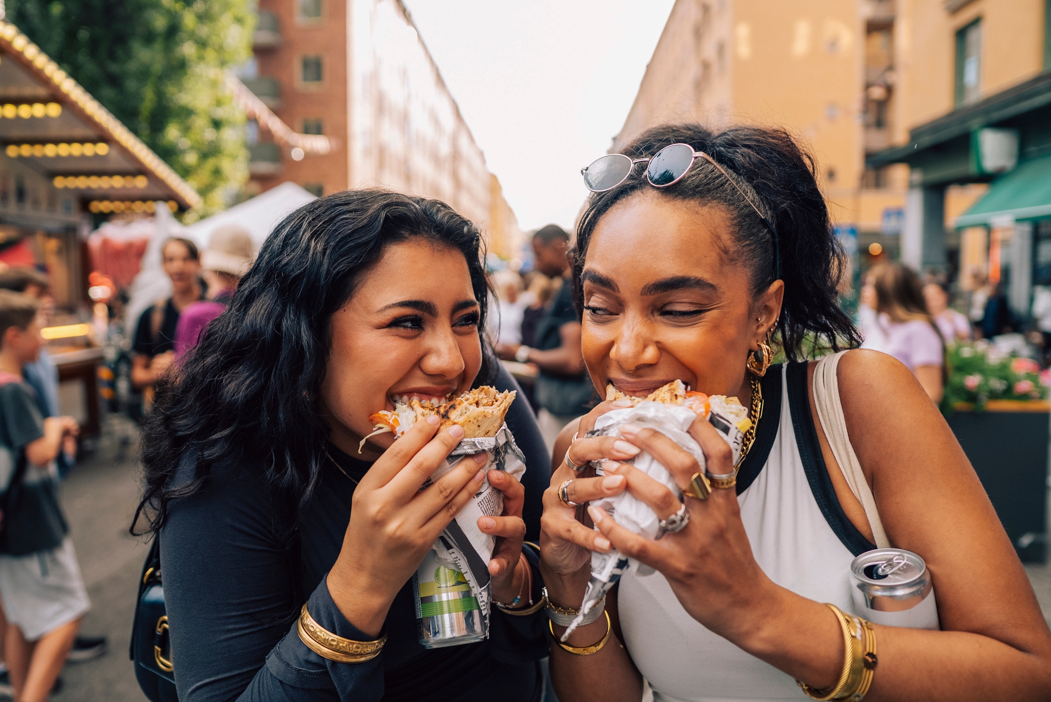 Two people enjoying sandwiches at an outdoor market, laughing and holding drinks, with others and stalls in the background