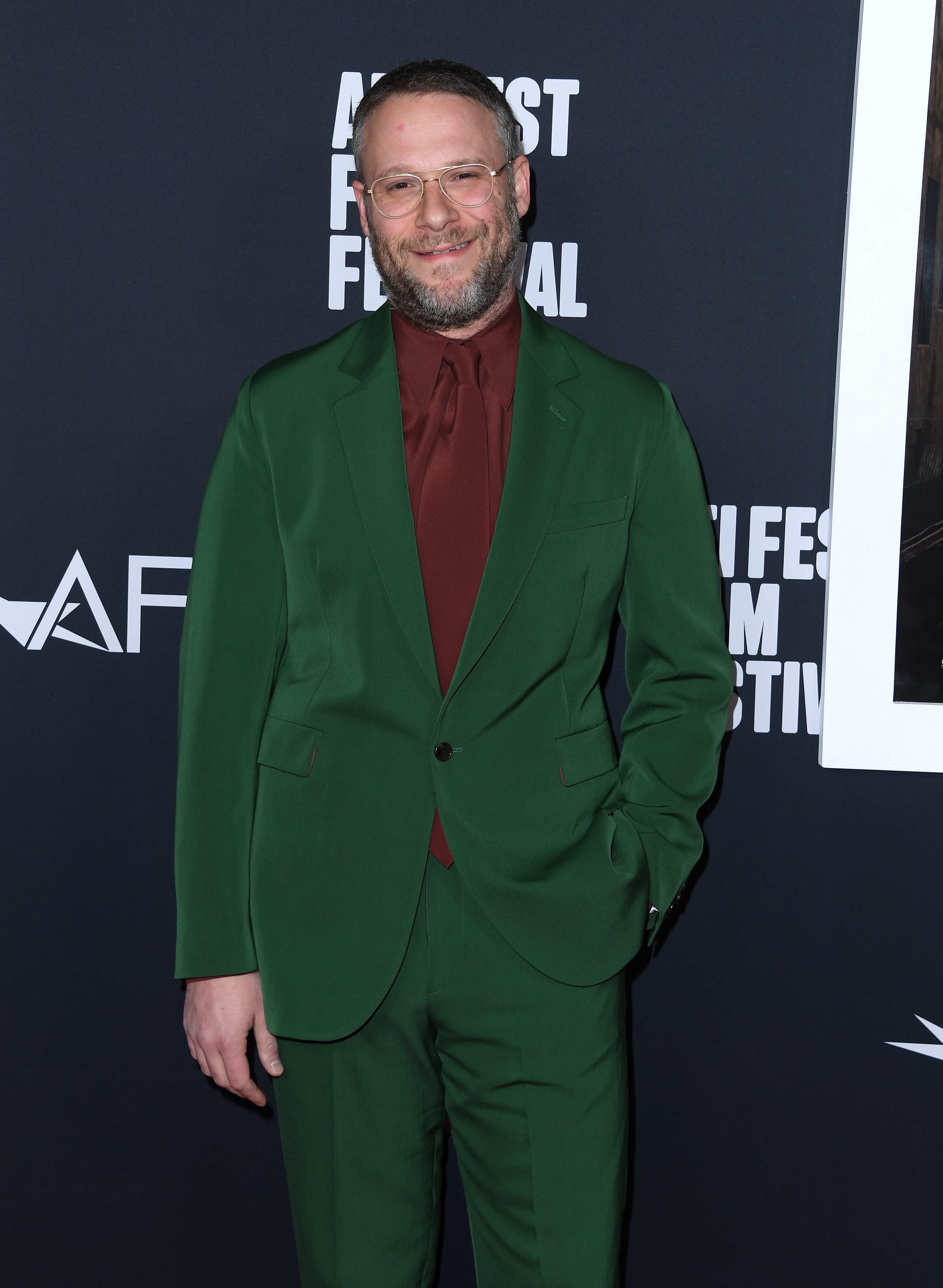 A idiosyncratic   connected  a reddish  carpet wears a tailored greenish  suit   with a burgundy shirt, smiling and posing for photos