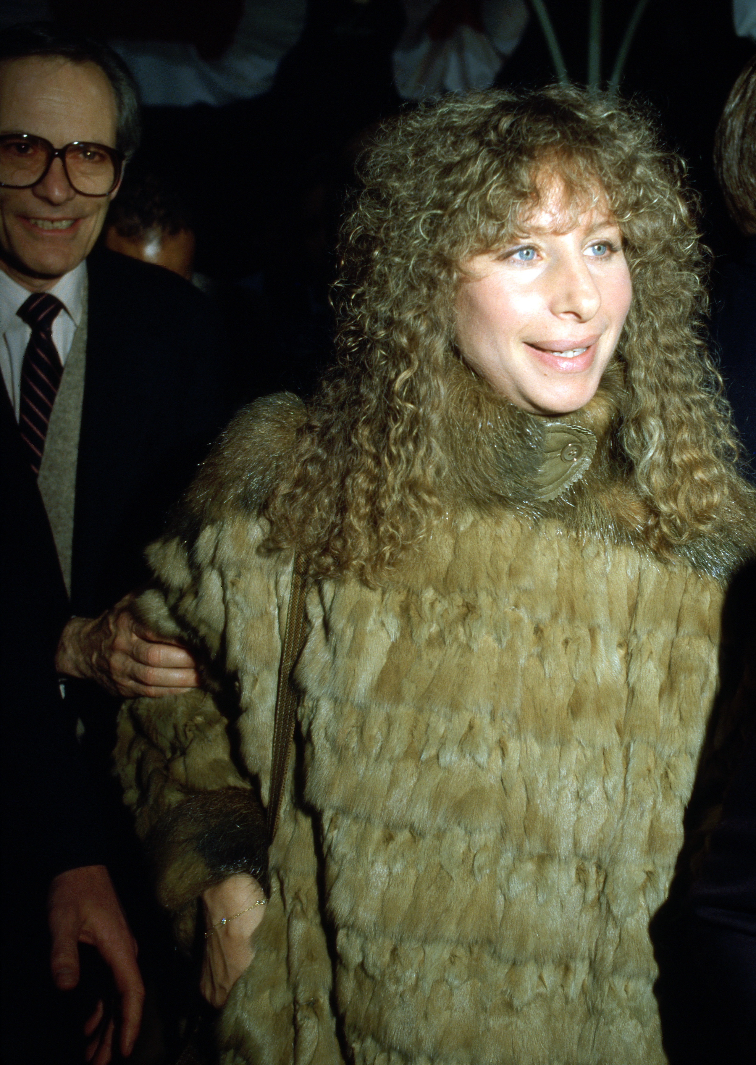 A pistillate   successful  a fur overgarment  smiles portion    lasting  connected  a reddish  carpet, accompanied by a antheral   successful  a suit   and necktie  successful  the background