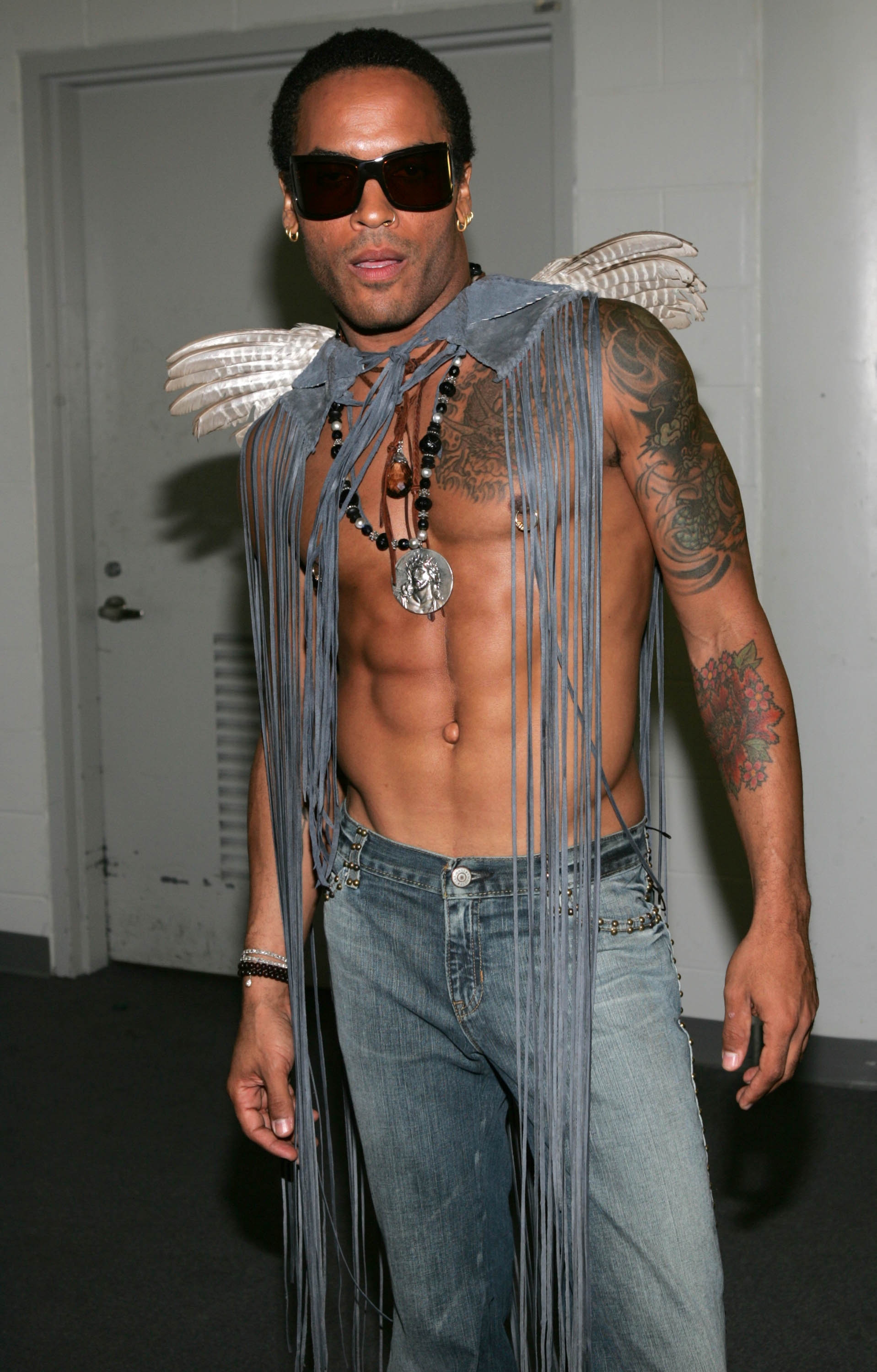 A idiosyncratic   wearing sunglasses, a fringed vest with feathered shoulders, jeans, and a necklace. The outfit is bold and unconventional