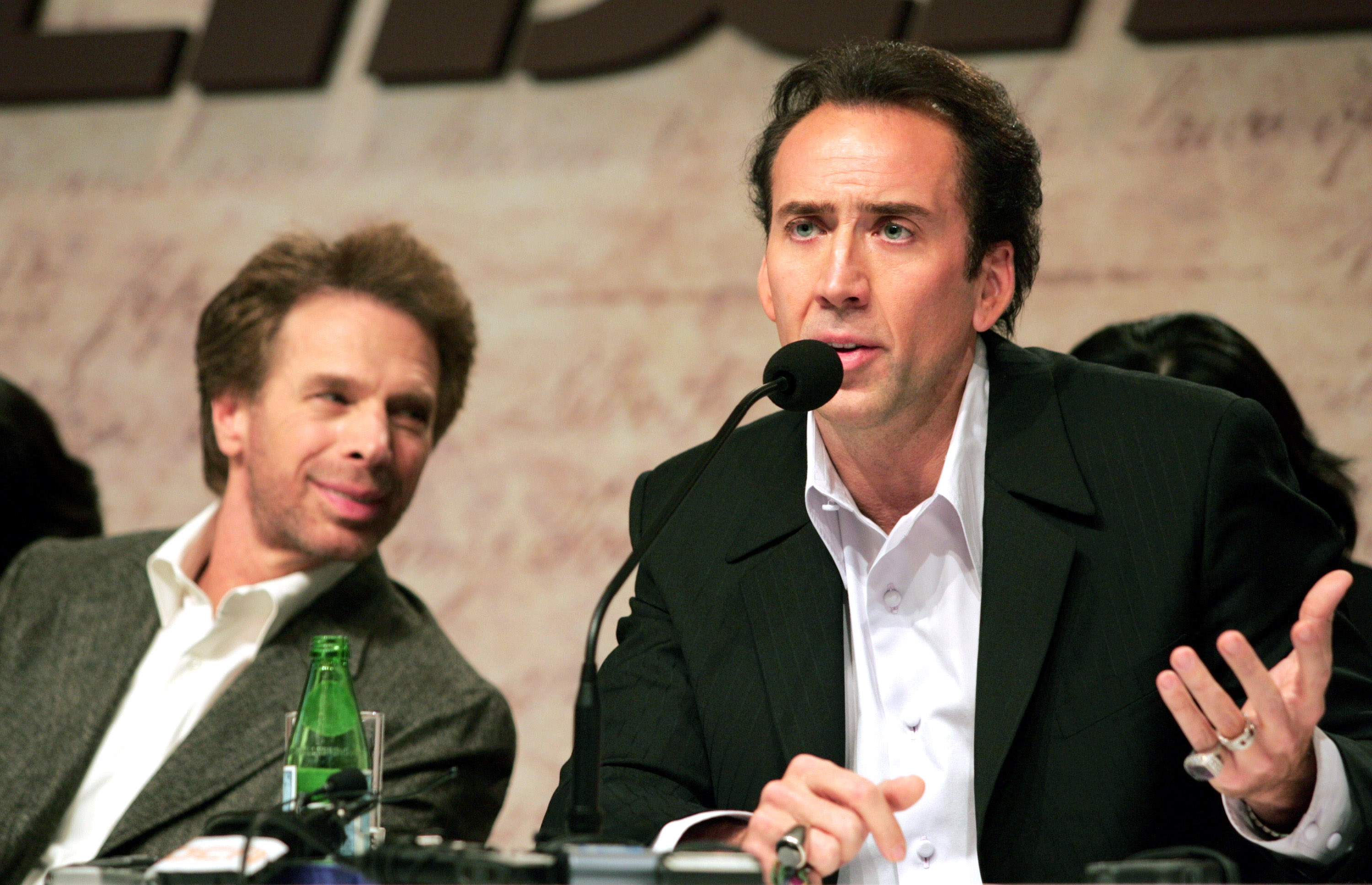 Two men sitting astatine  a property   lawsuit   table. One antheral   speaks into a microphone, wearing a suit, the different   listens attentively with a flimsy  smile