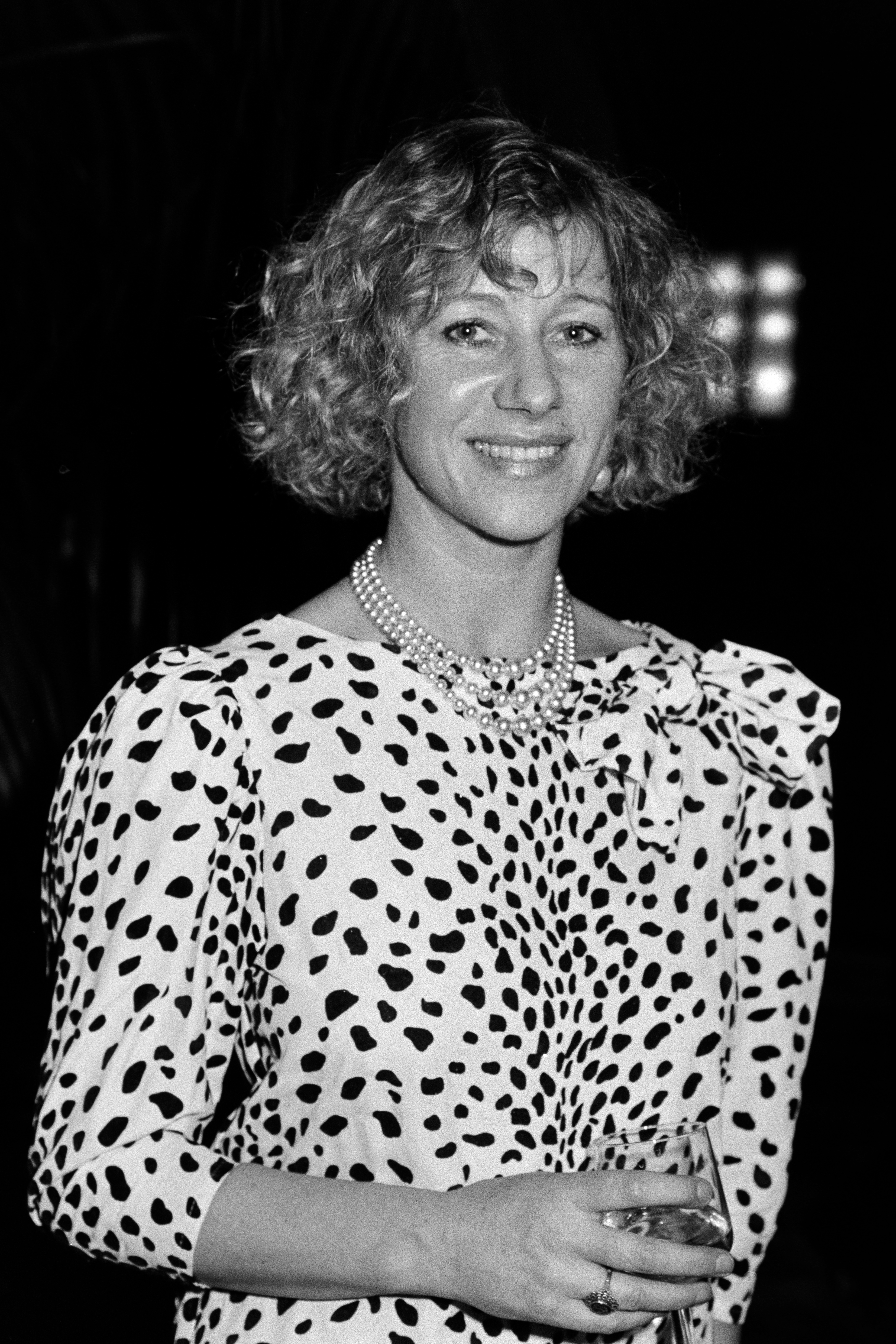 Person wearing a polka dot formal  with puffed sleeves and a pearl necklace, holding a glass, smiling astatine  an event