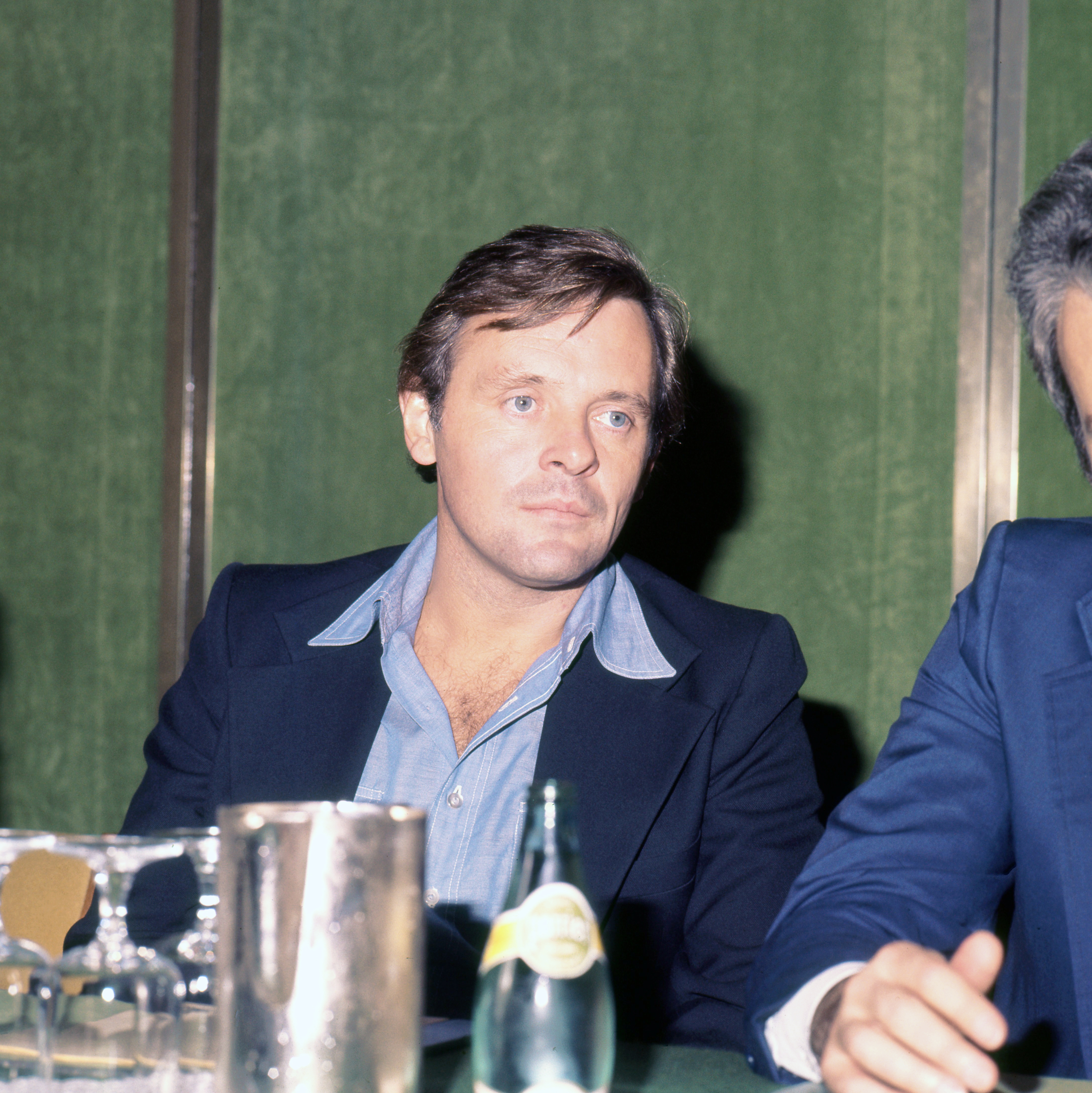 A antheral   successful  a casual suit   sits astatine  a array  with drinks and glasses, looking ahead. Another idiosyncratic   is partially disposable   beside him