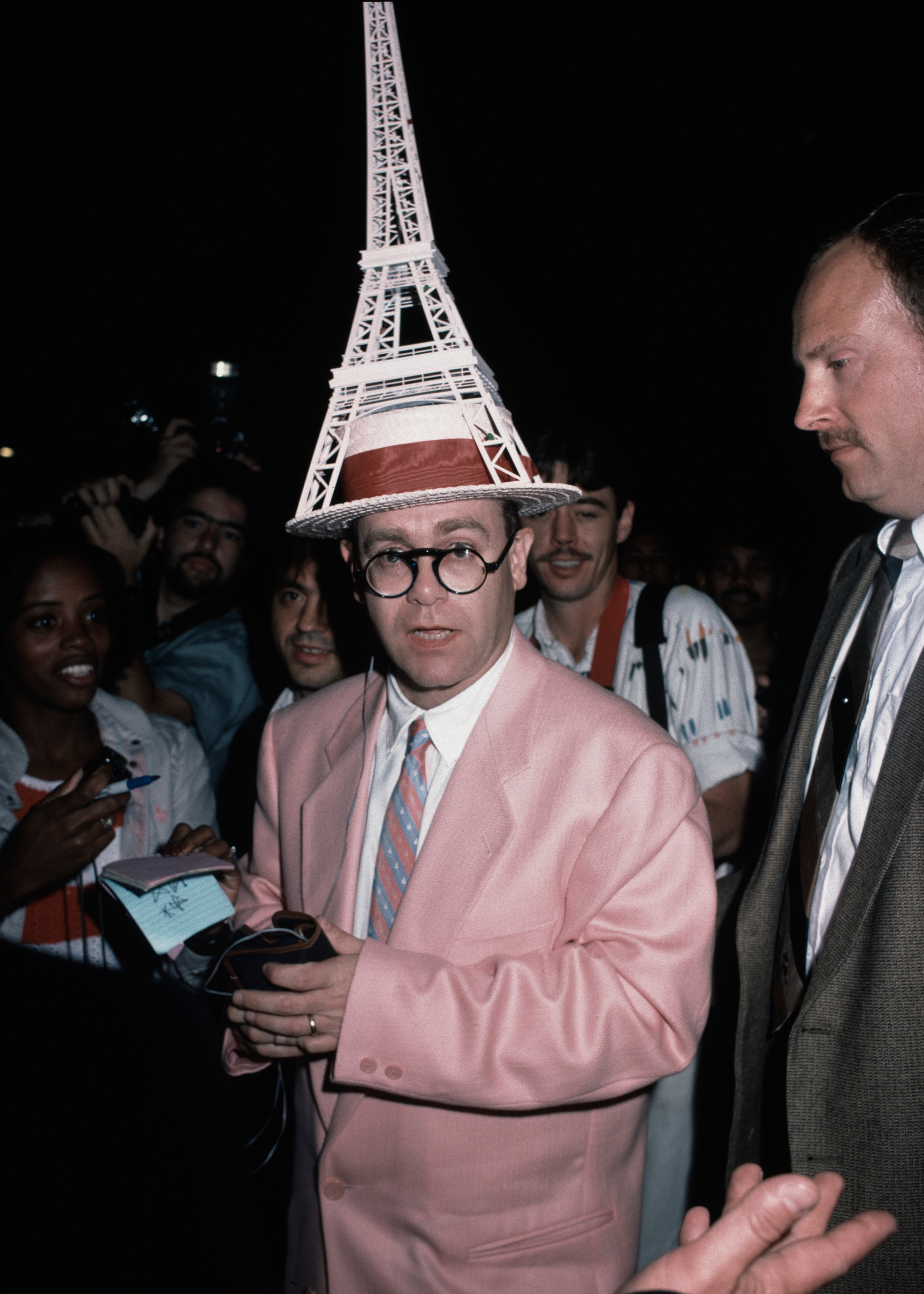 Person successful  a whimsical Eiffel Tower chapeau  and pinkish  suit, surrounded by fans and information    astatine  a personage  event